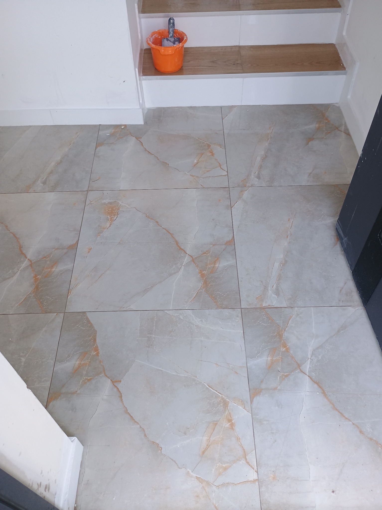 Wykończona podłoga z płytek imitujących marmur z pomarańczowym żyłkowaniem, widoczny fragment schodów z drewnopodobnymi panelami i pomarańczowe wiadro z narzędziami glazurniczymi.