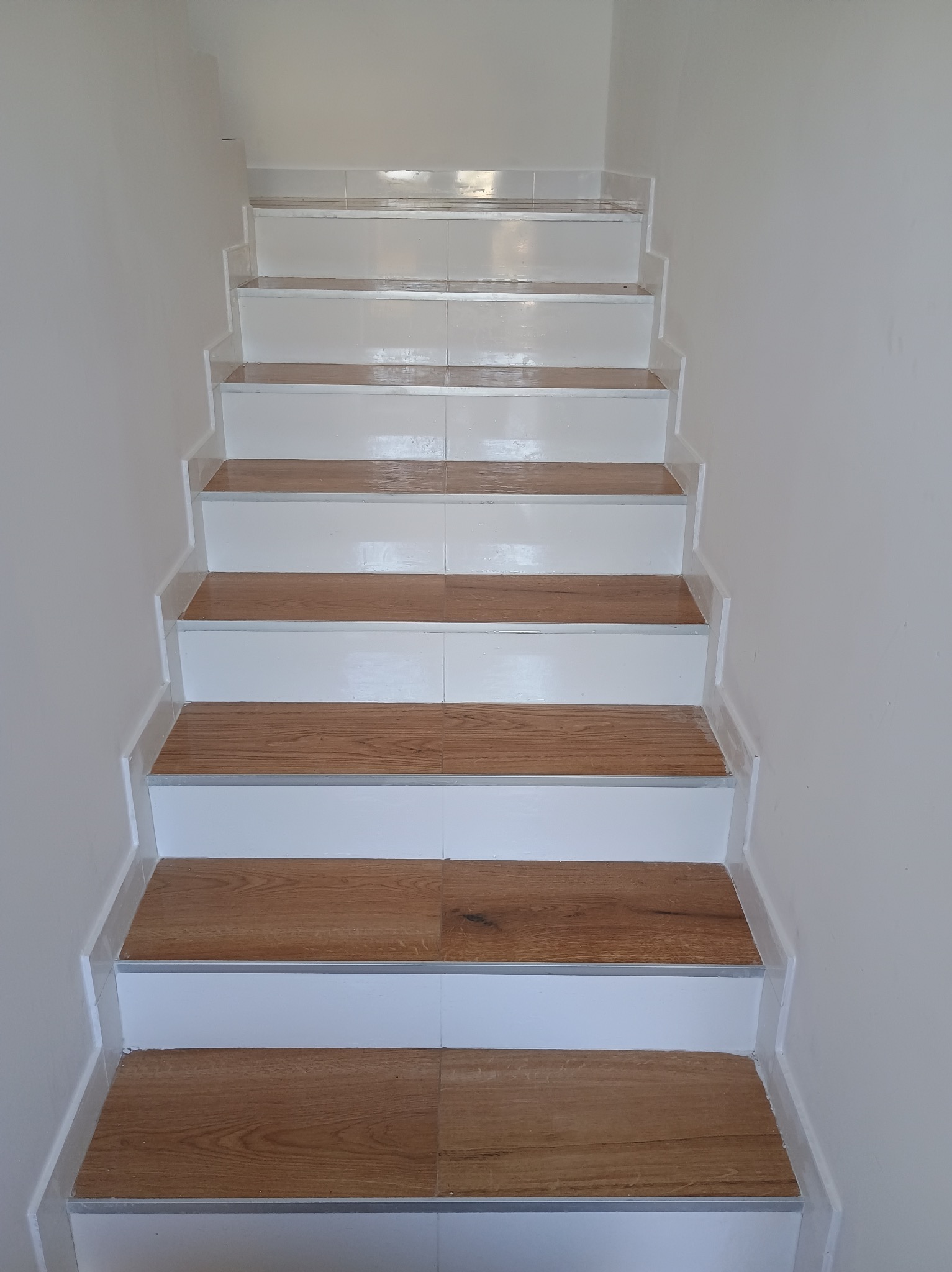 Schody wewnętrzne z białymi stopnicami i okładziną imitującą drewno, wykończone listwami przypodłogowymi.