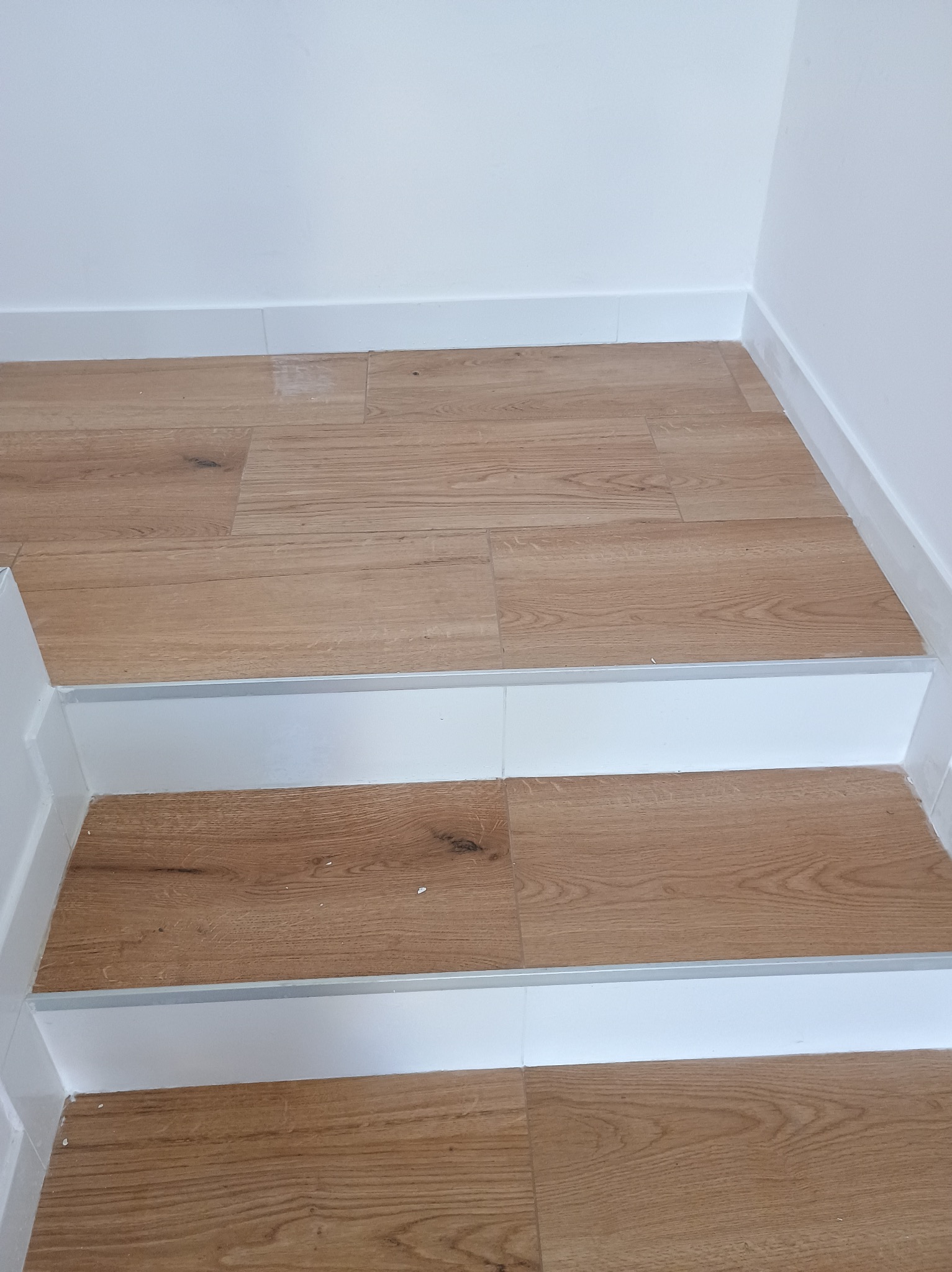 Wykończone schody z drewnopodobnych paneli z białymi listwami przypodłogowymi i aluminiowymi profilami krawędziowymi.