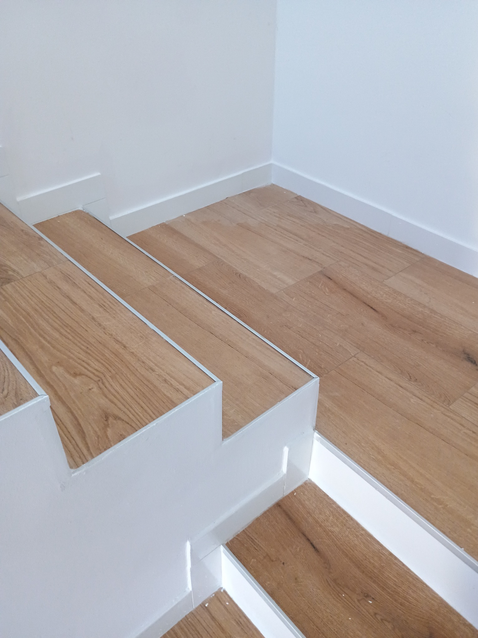 Schody z drewnopodobnymi panelami i białymi listwami przypodłogowymi, wykończone srebrnymi profilami na krawędziach stopni.