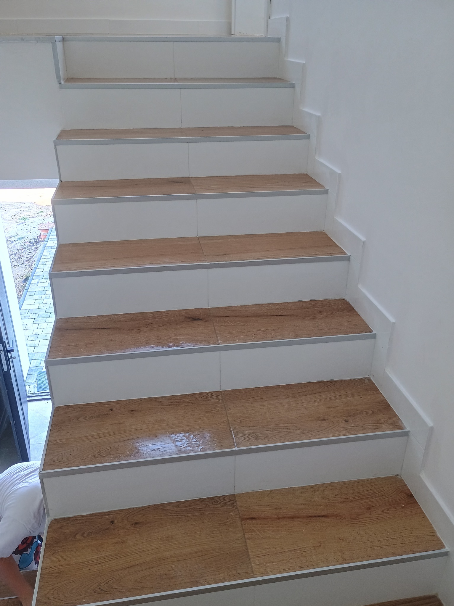 Wykończone schody z drewnopodobnymi płytkami, białe stopnice z szarą listwą wykończeniową, widoczny fragment osoby w białej koszulce.