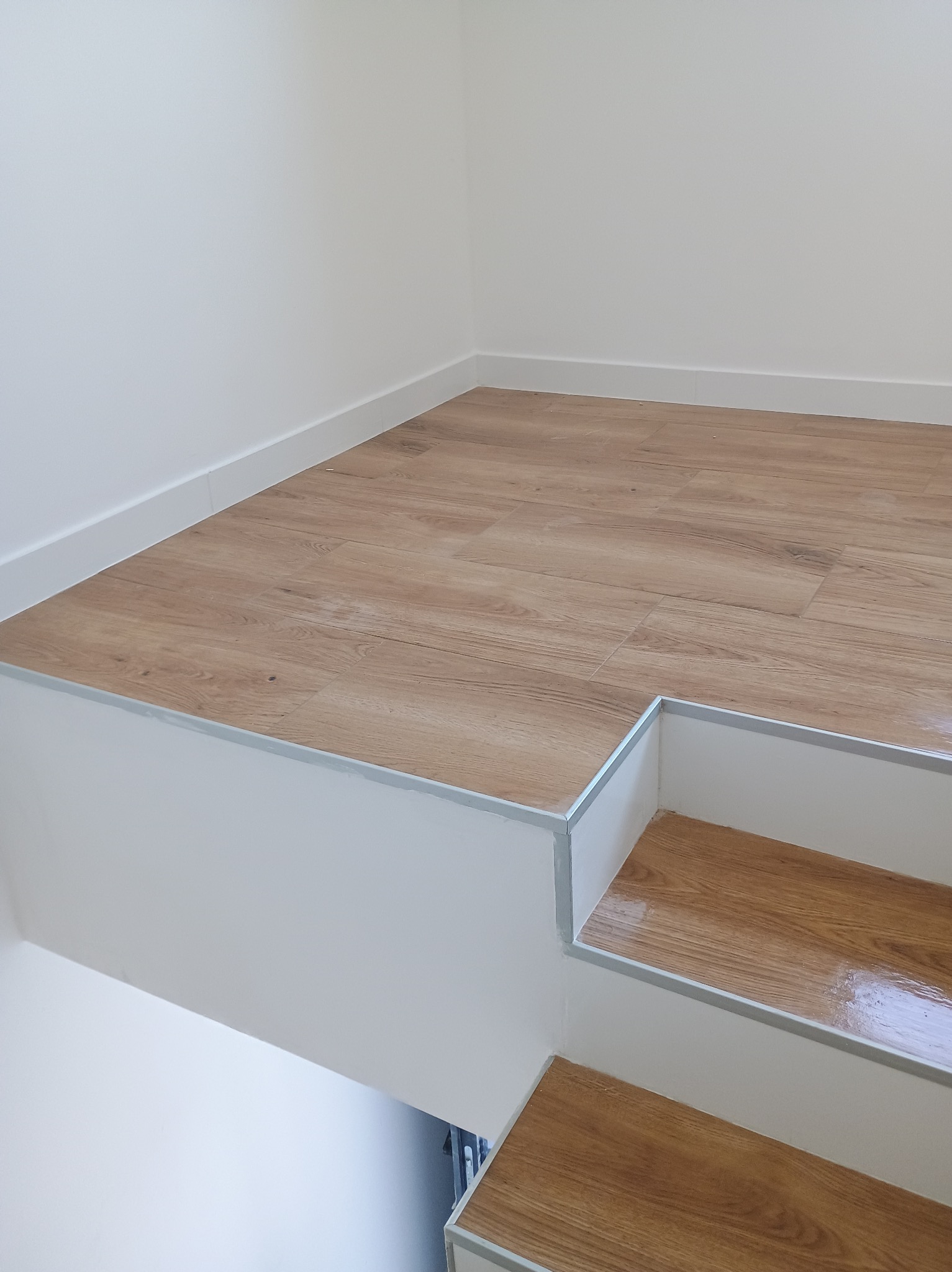 Jasne panele podłogowe imitujące drewno na podwyższeniu z dwoma stopniami, wykończone srebrnymi listwami, białe ściany.