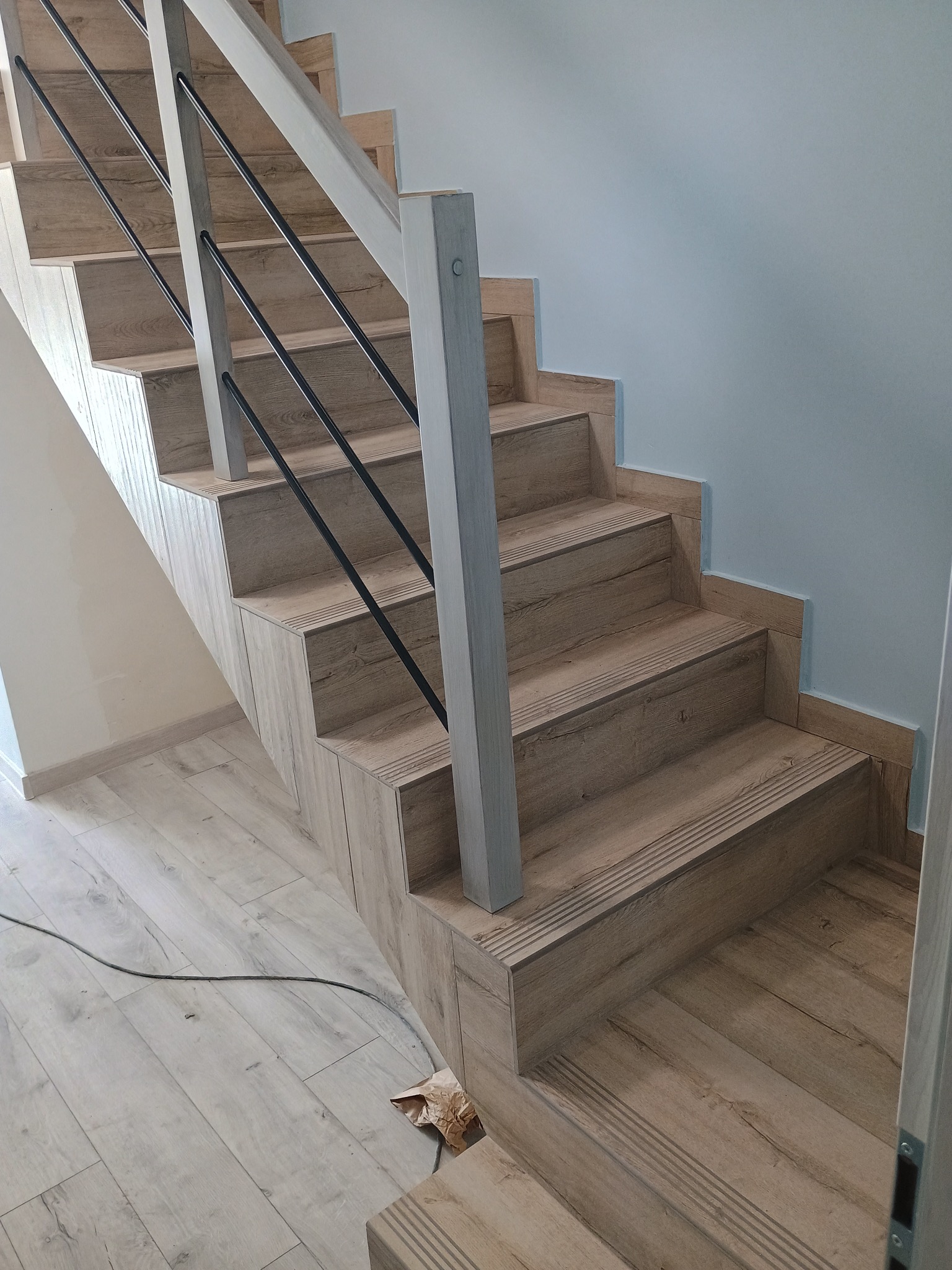 Schody wewnętrzne wykończone panelami drewnopodobnymi, z czarną metalową balustradą i jasnoszarymi słupkami, dopasowane do podłogi w odcieniach bieli i szarości.