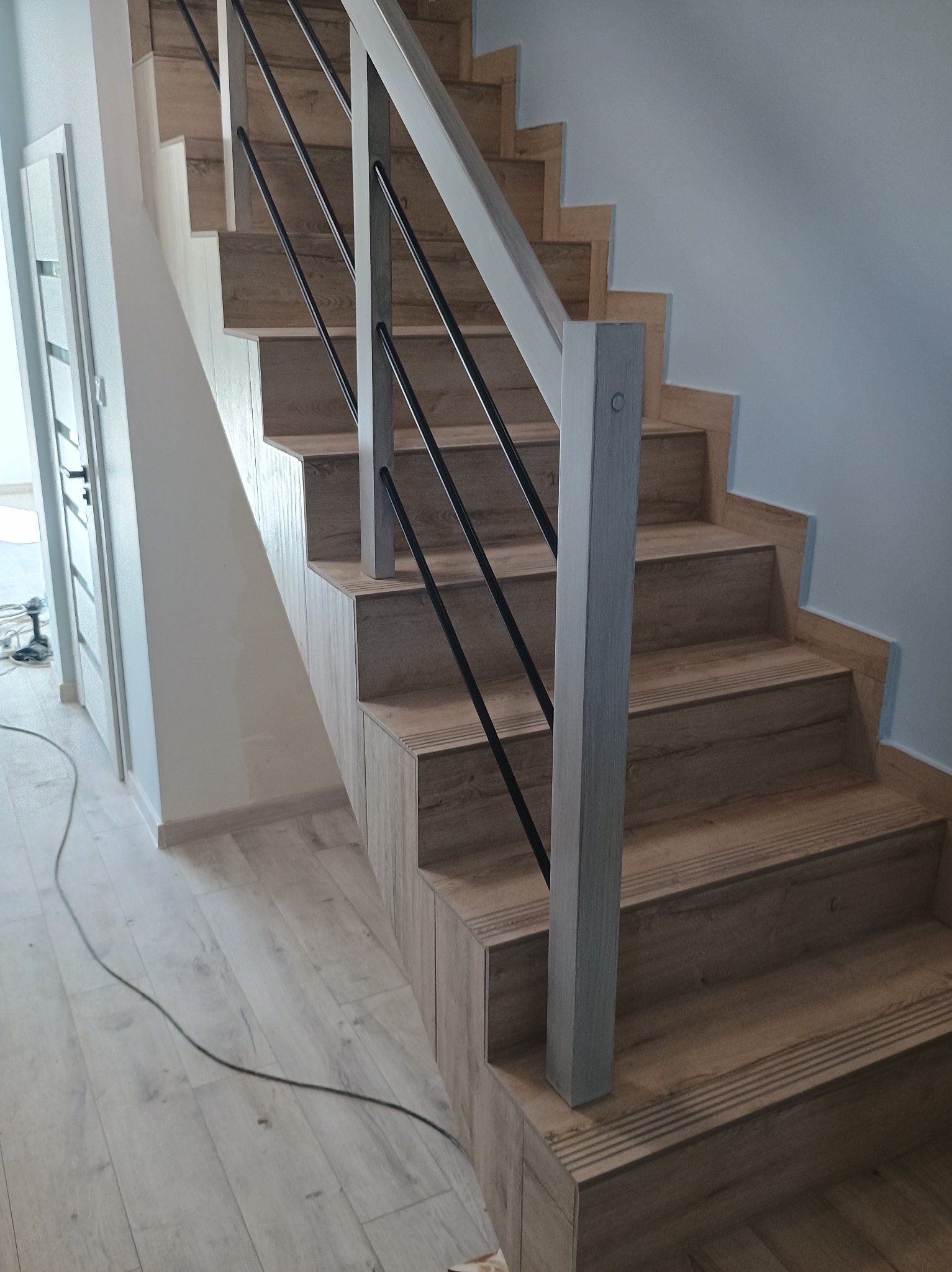 Wykończone schody z drewnopodobnych płytek z czarnymi metalowymi balustradami i jasnymi drewnianymi poręczami, widoczne narzędzia i kable w tle.