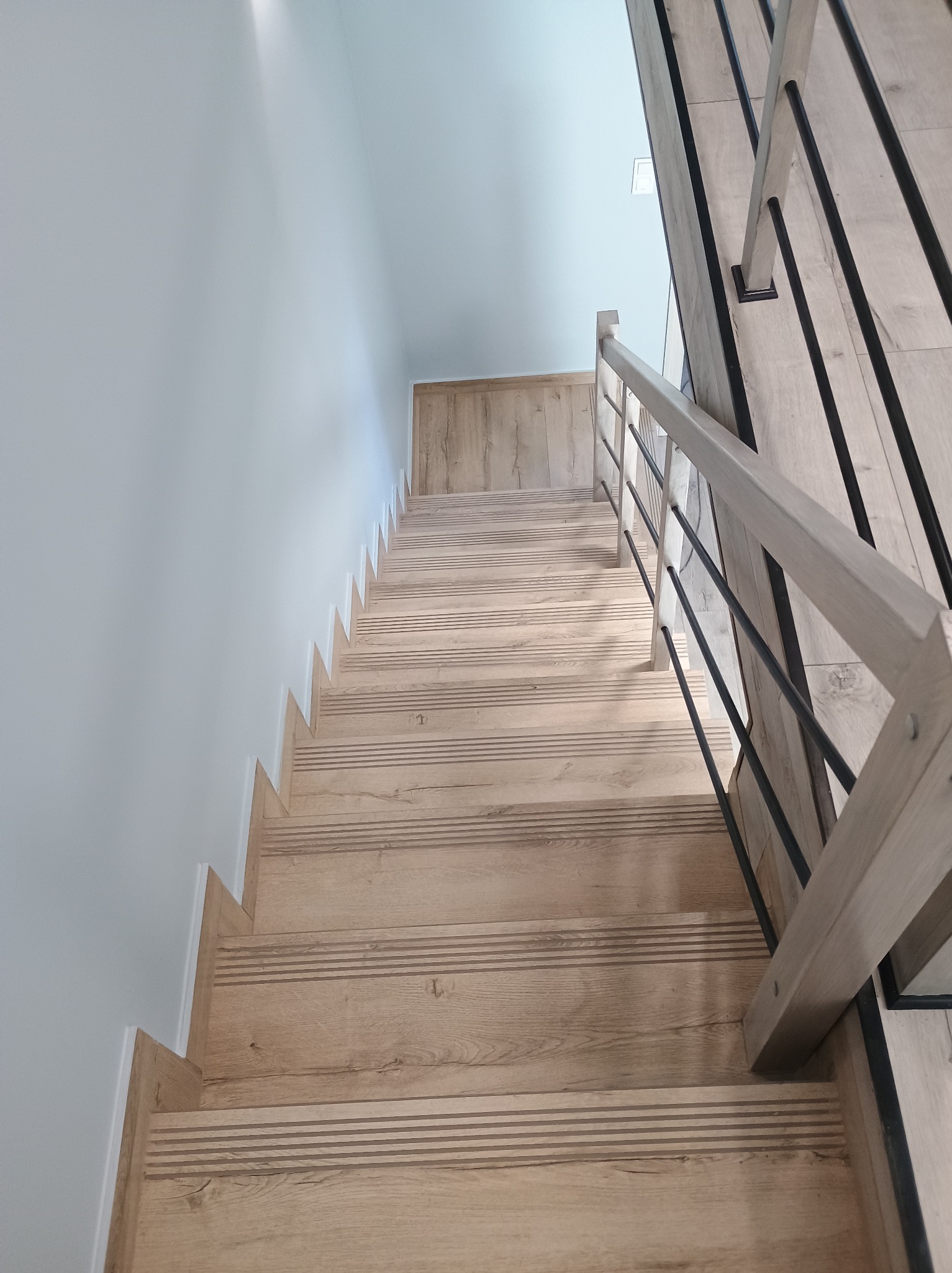 Schody drewniane z jasnego drewna z poziomymi rowkami, białe ściany i czarne metalowe balustrady, widok z góry.