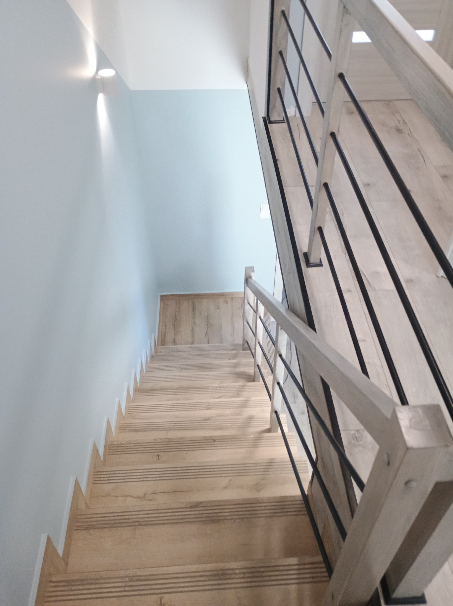 Jasne, drewniane schody z czarnymi, metalowymi balustradami w nowoczesnym wnętrzu, oświetlone kinkietem na ścianie.
