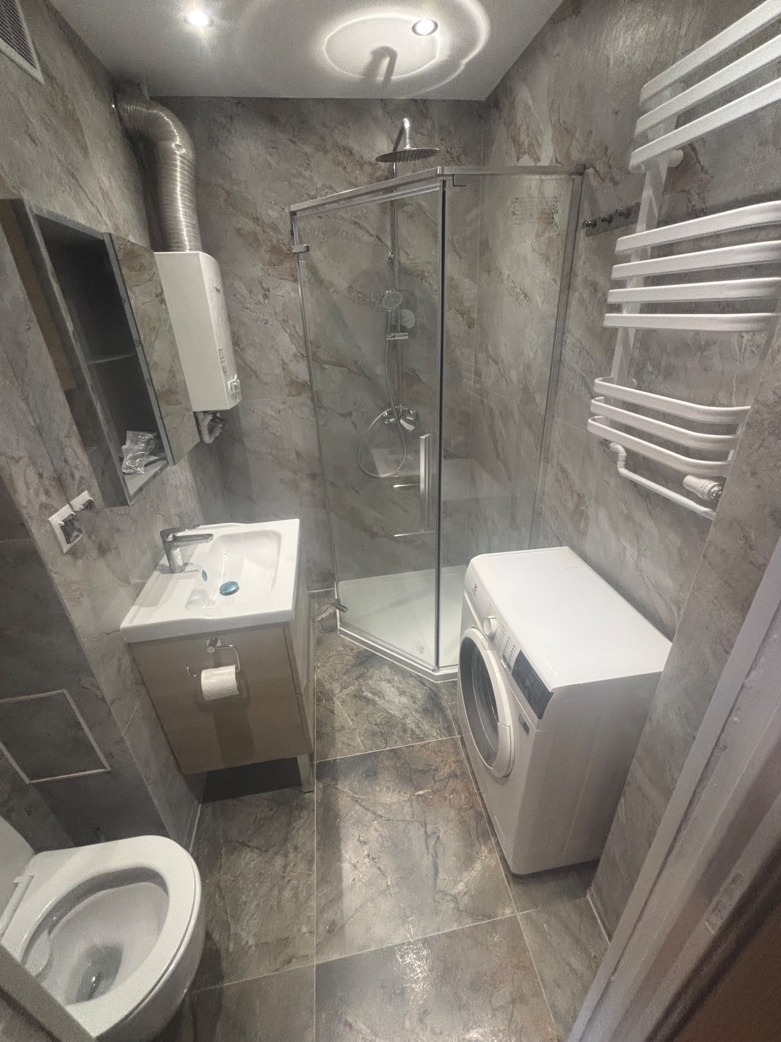 Wykończona łazienka w szarościach: kabina prysznicowa, umywalka z szafką, toaleta, pralka i grzejnik drabinkowy na tle marmurowych płytek.