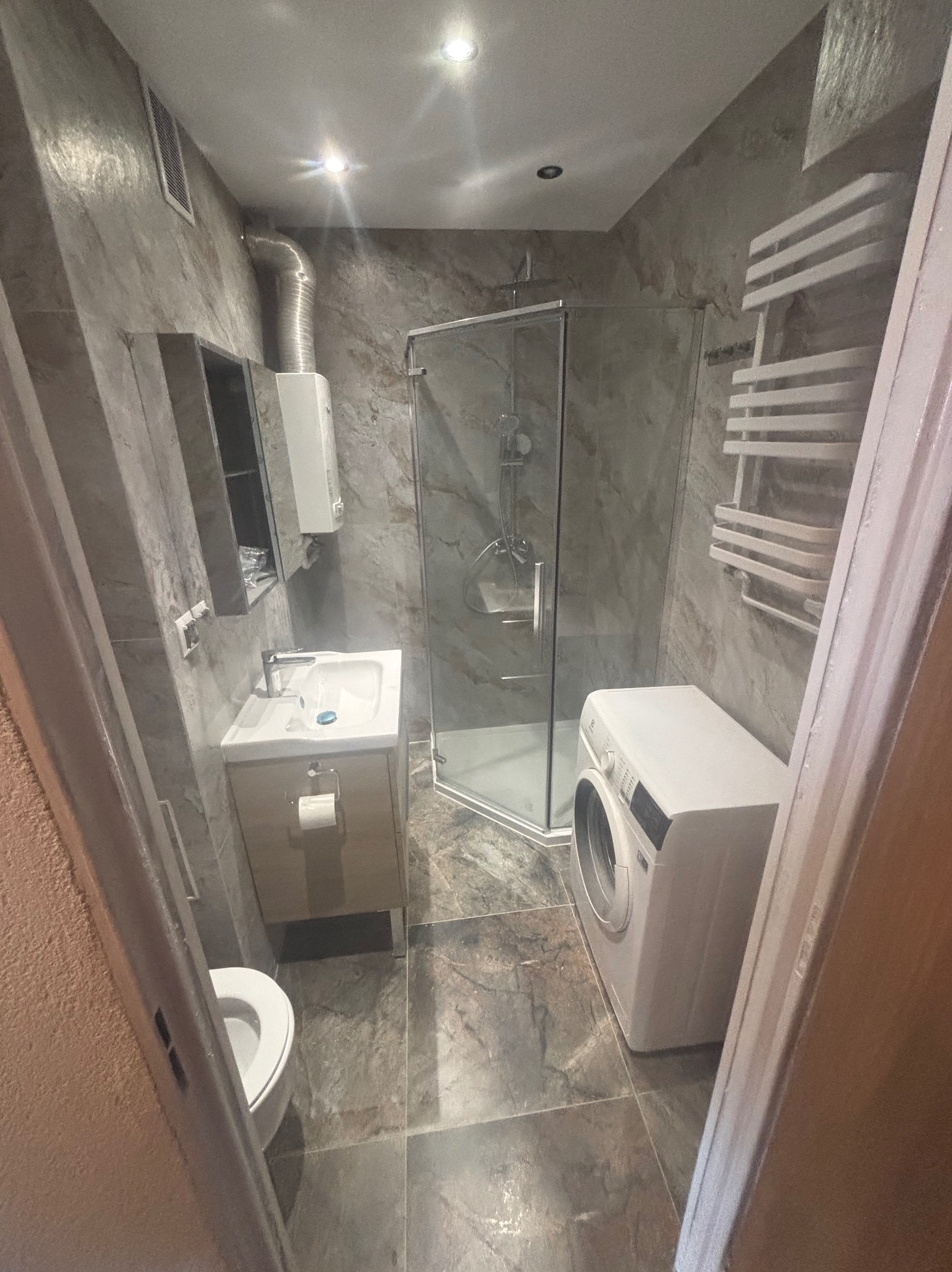 Nowoczesna łazienka po remoncie: kabina prysznicowa, umywalka z szafką, toaleta i pralka. Wykończenie w odcieniach szarości z marmurkową teksturą na ścianach.