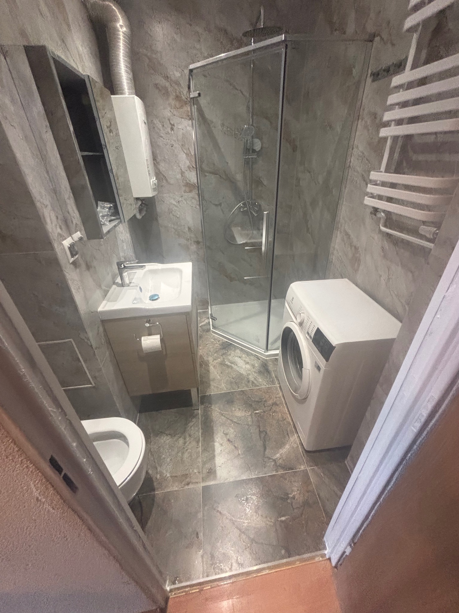 Wykończona łazienka w szarościach: kabina prysznicowa, umywalka z szafką, toaleta i pralka. Na ścianach płytki imitujące marmur, grzejnik drabinkowy.