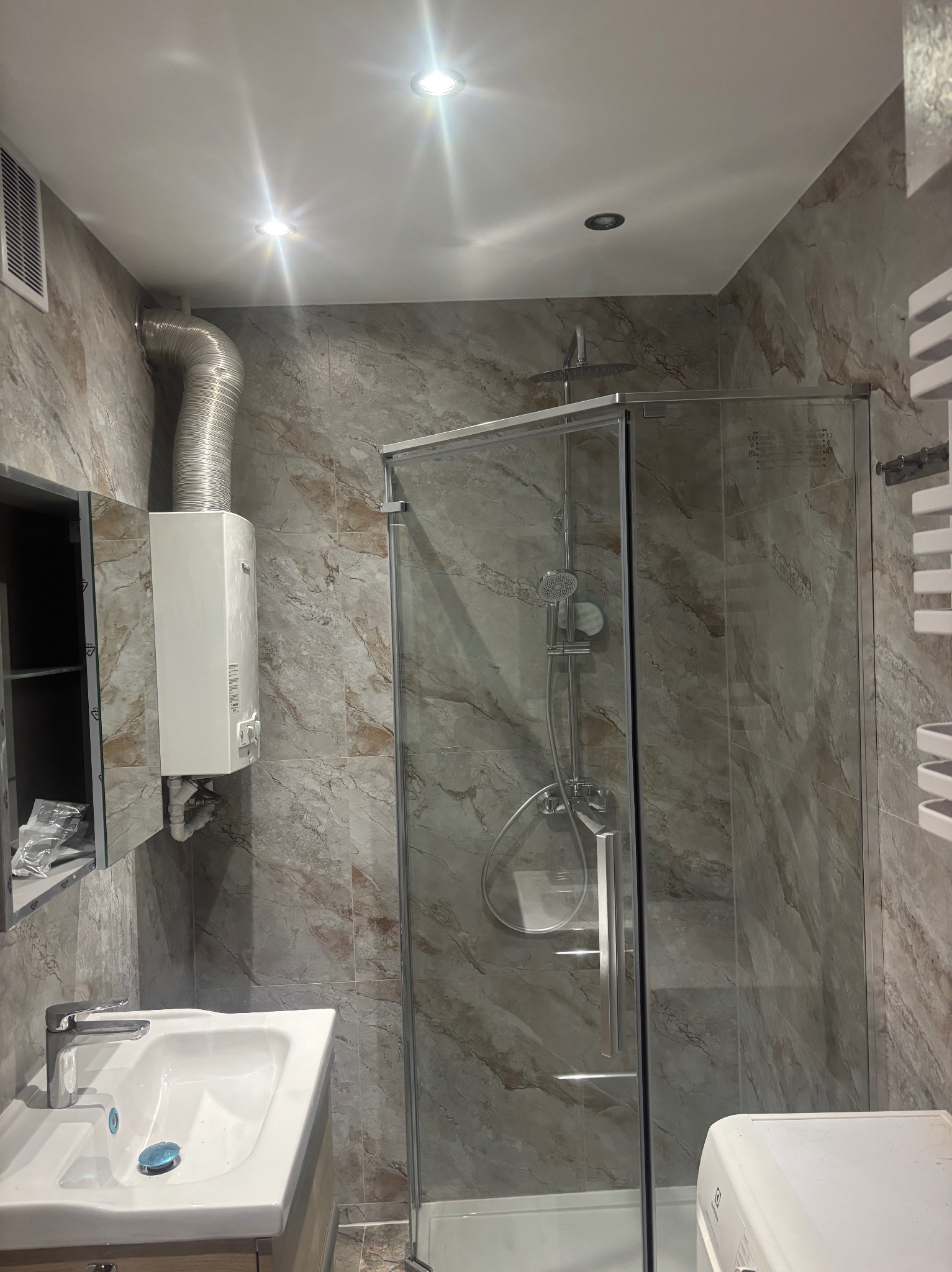 Wykończona łazienka z kabiną prysznicową, umywalką i grzejnikiem drabinkowym. Płytki imitujące marmur na ścianach. Zamknięta szafka z lustrem i wiszący piec gazowy.