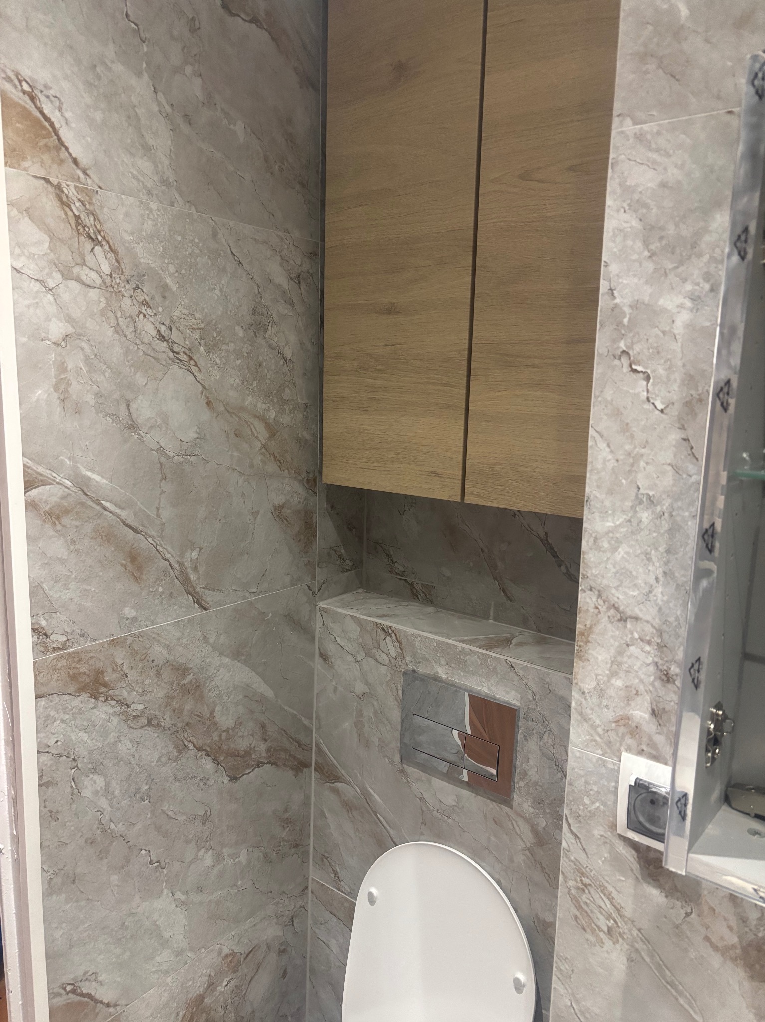 Wykończona łazienka z marmurowymi płytkami, zabudową nad toaletą i lustrem. Uwagę zwraca elegancki przycisk spłuczki w kolorze miedzi.