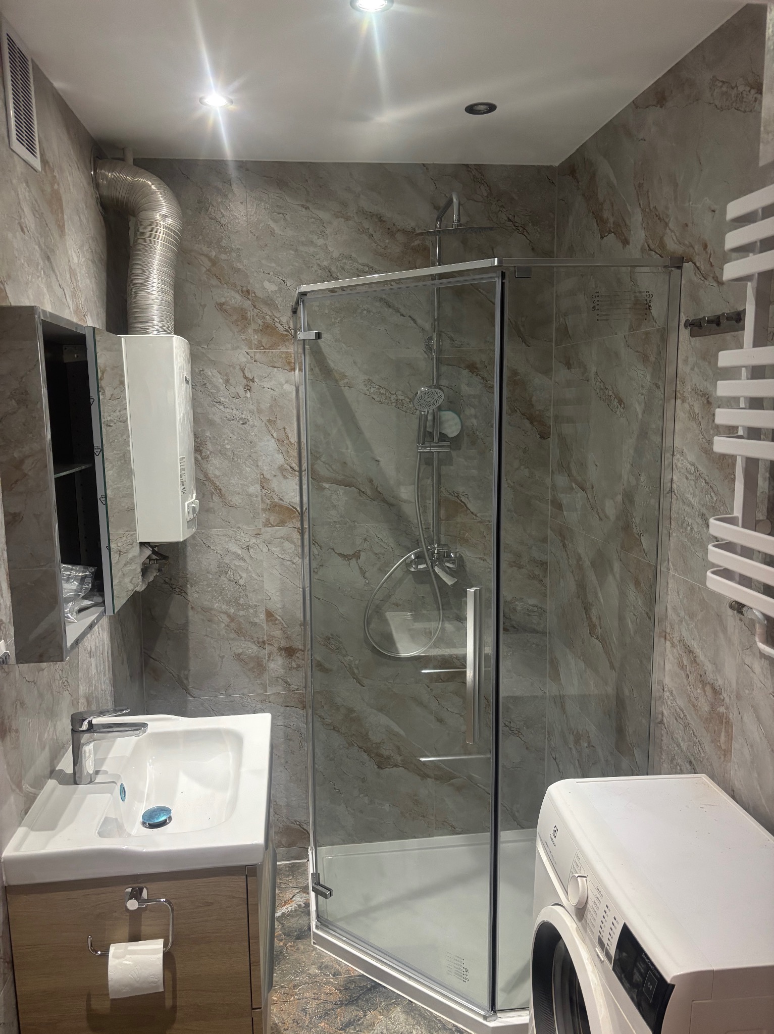 Wykończona łazienka z kabiną prysznicową, umywalką z szafką, pralką i grzejnikiem drabinkowym na ścianie z płytkami imitującymi marmur.