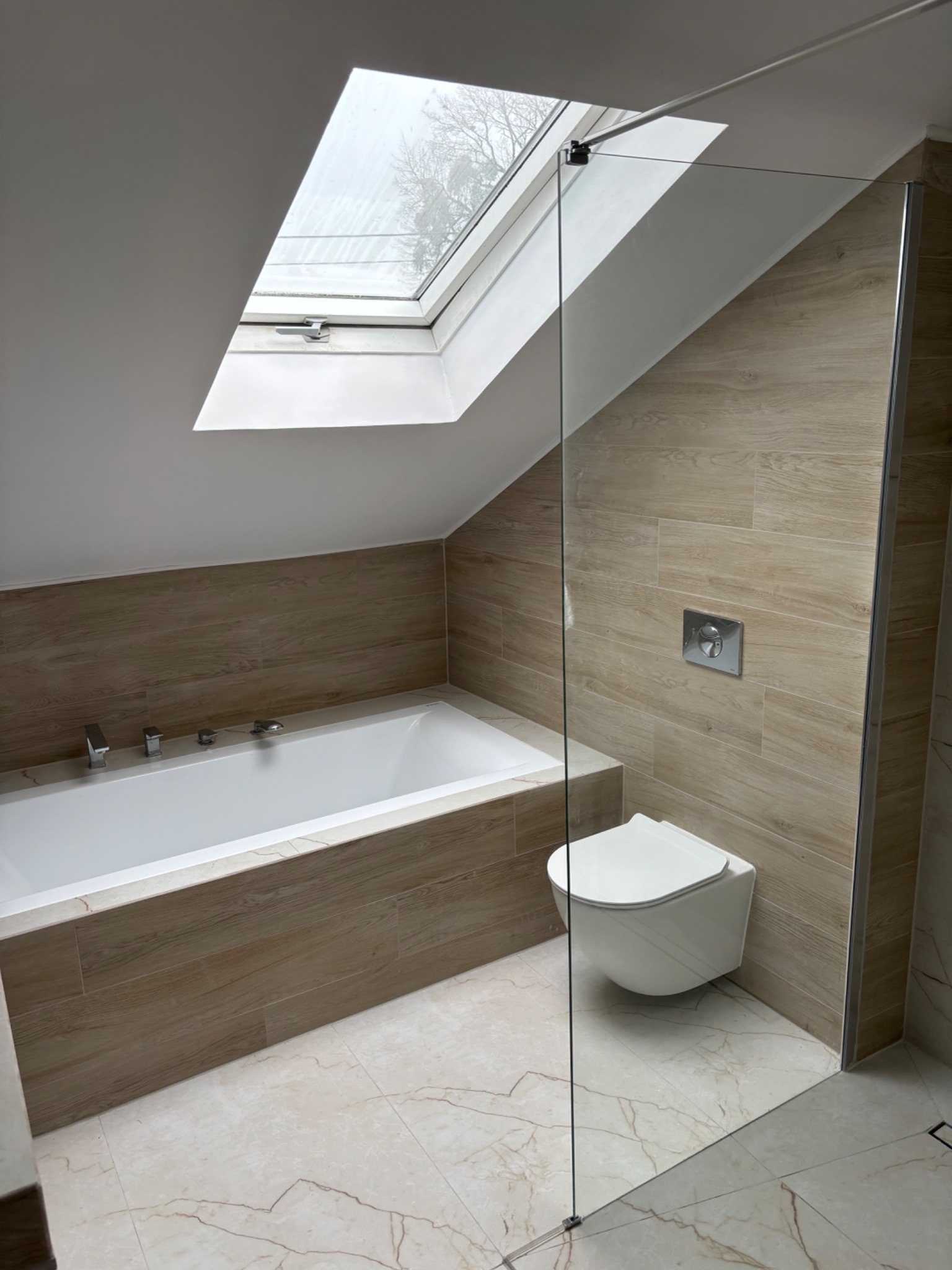 Nowoczesna łazienka na poddaszu z oknem dachowym, beżowe płytki imitujące drewno, biała wanna i toaleta podwieszana, szklana kabina prysznicowa.