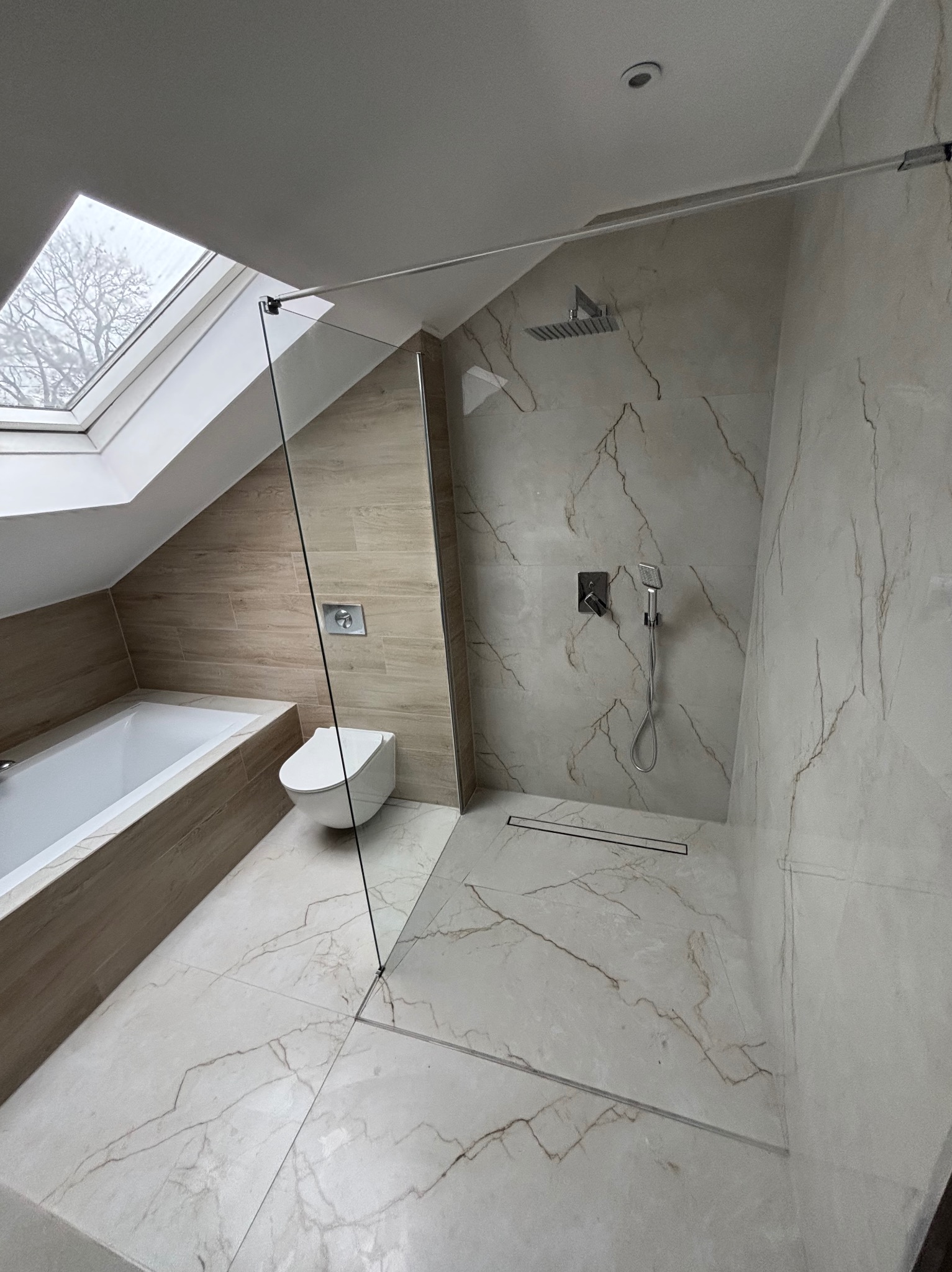 Nowoczesna łazienka na poddaszu z kabiną prysznicową typu walk-in, beżowymi płytkami imitującymi marmur i oknem dachowym. Minimalistyczna armatura i odpływ liniowy.