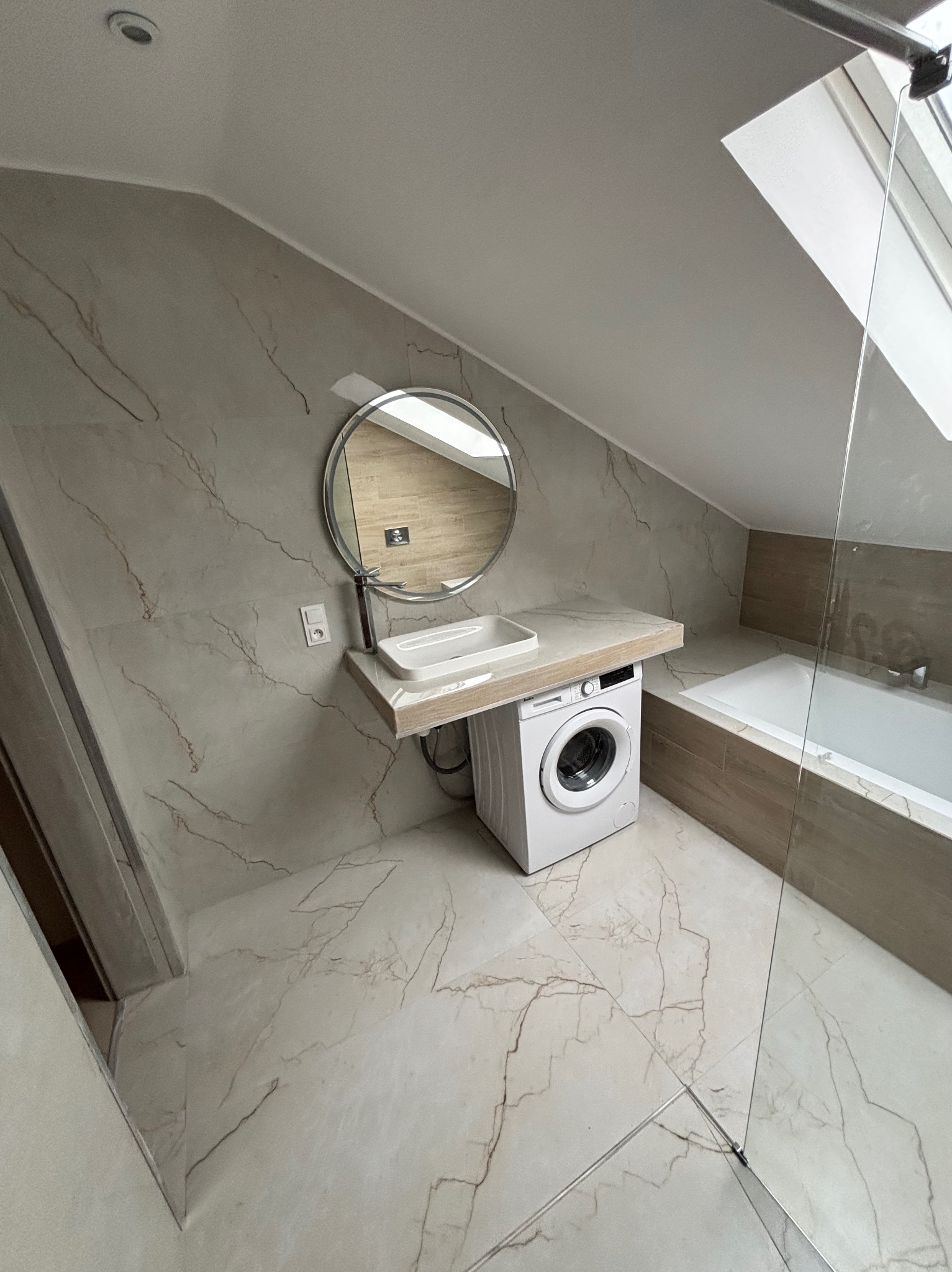 Stylowa łazienka na poddaszu w Żorach z marmurowymi płytkami, okrągłym lustrem nad umywalką i pralką pod blatem. Kabina prysznicowa ze szklaną ścianką.