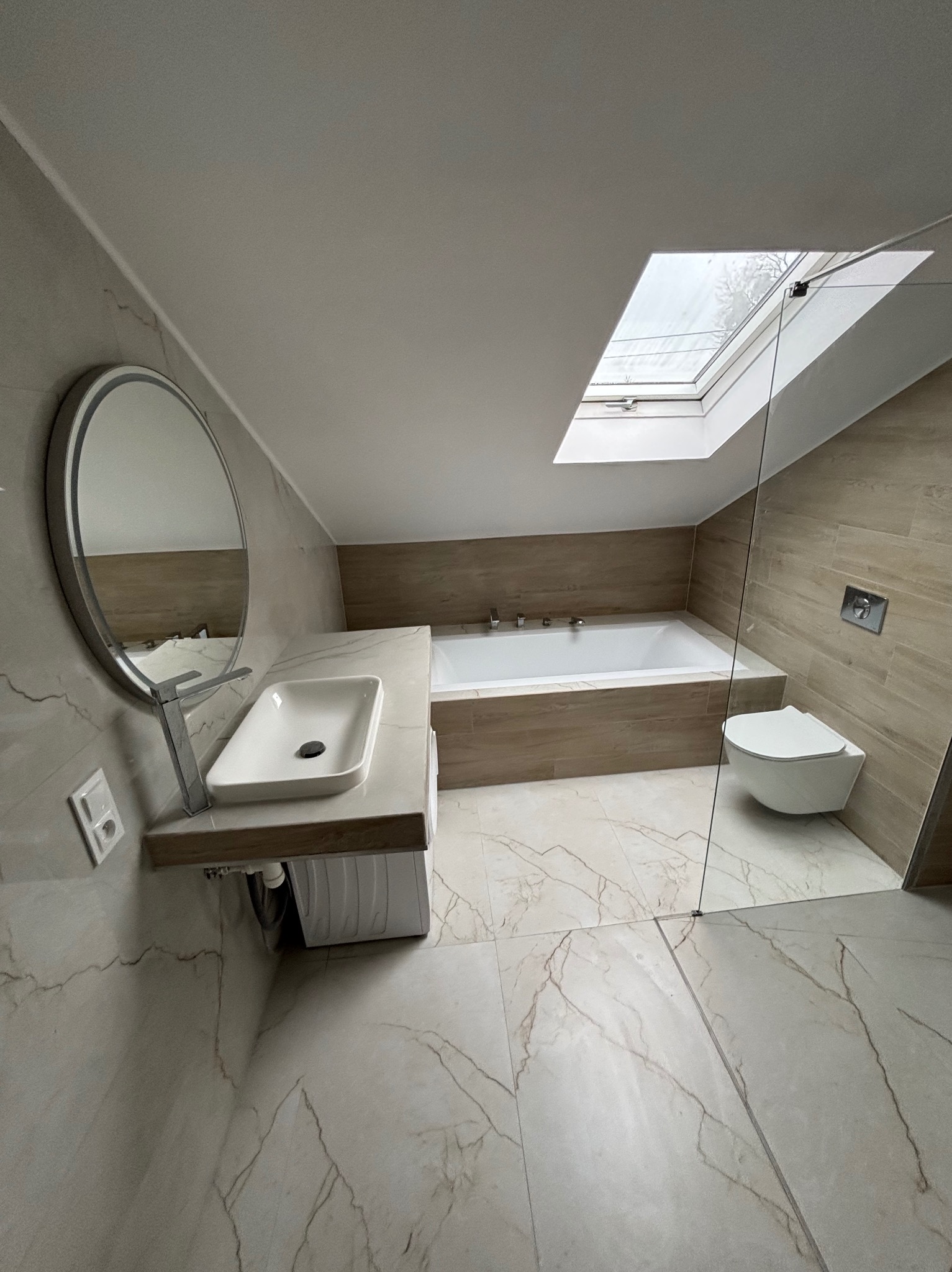 Nowoczesna łazienka na poddaszu z oknem dachowym. Umywalka nablatowa, owalne lustro, beżowe płytki imitujące drewno i marmurowa podłoga. Minimalistyczny design.