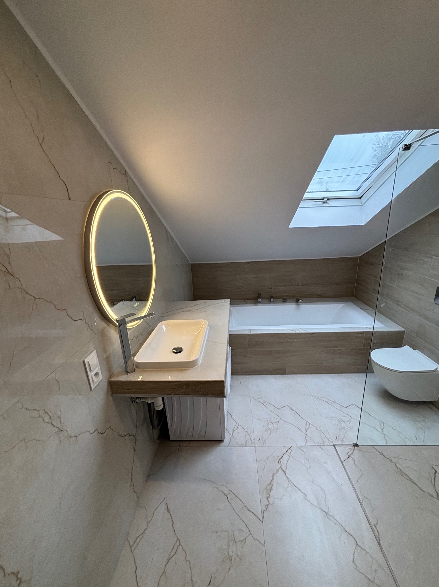 Jasna łazienka na poddaszu z oknem dachowym, marmurowymi płytkami, owalnym lustrem z podświetleniem LED i nowoczesną armaturą. Minimalistyczny design.