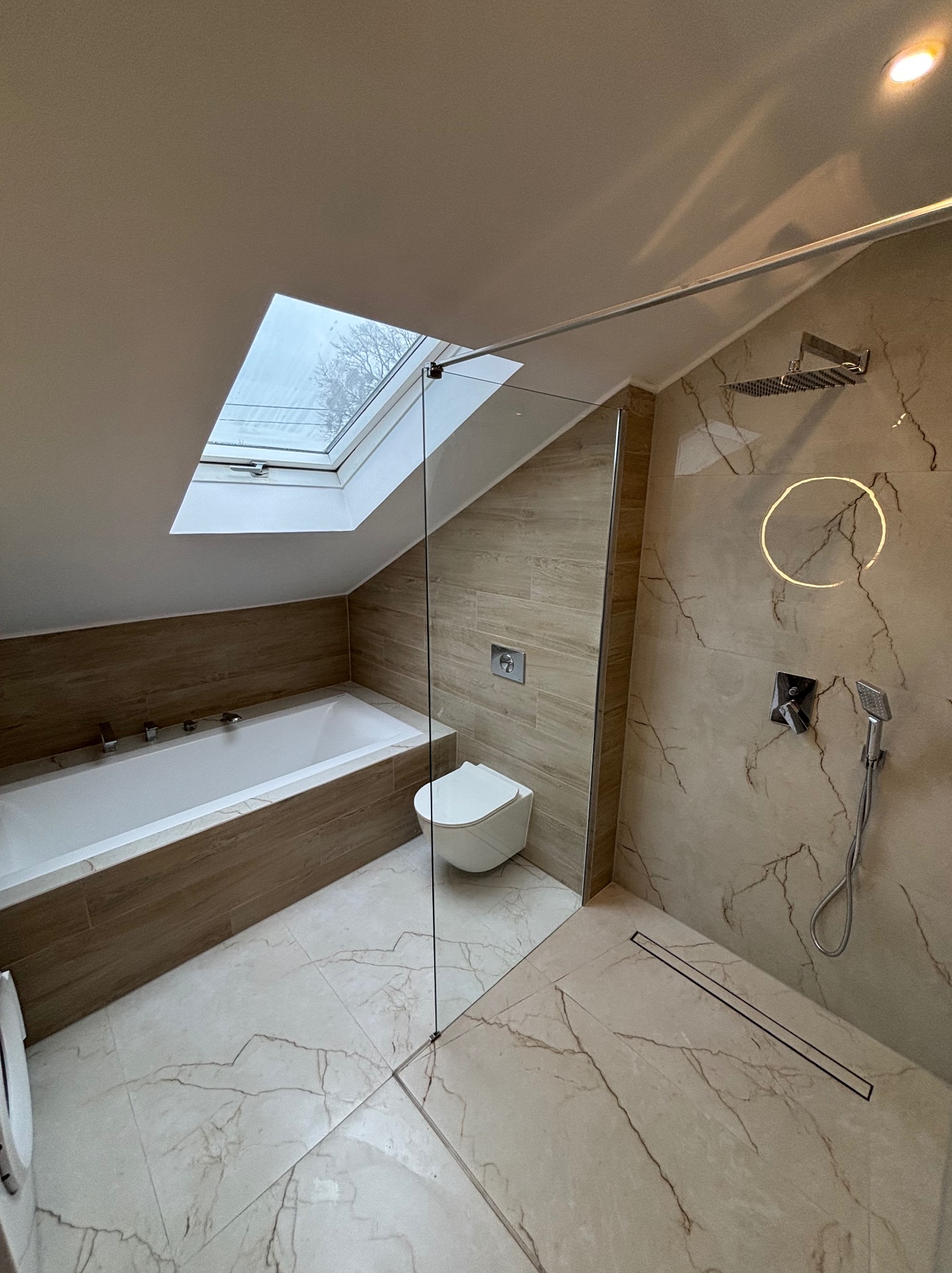 Jasna łazienka na poddaszu z wanną, toaletą, kabiną prysznicową typu walk-in i oknem dachowym. Beżowe płytki imitujące marmur na ścianach i podłodze.