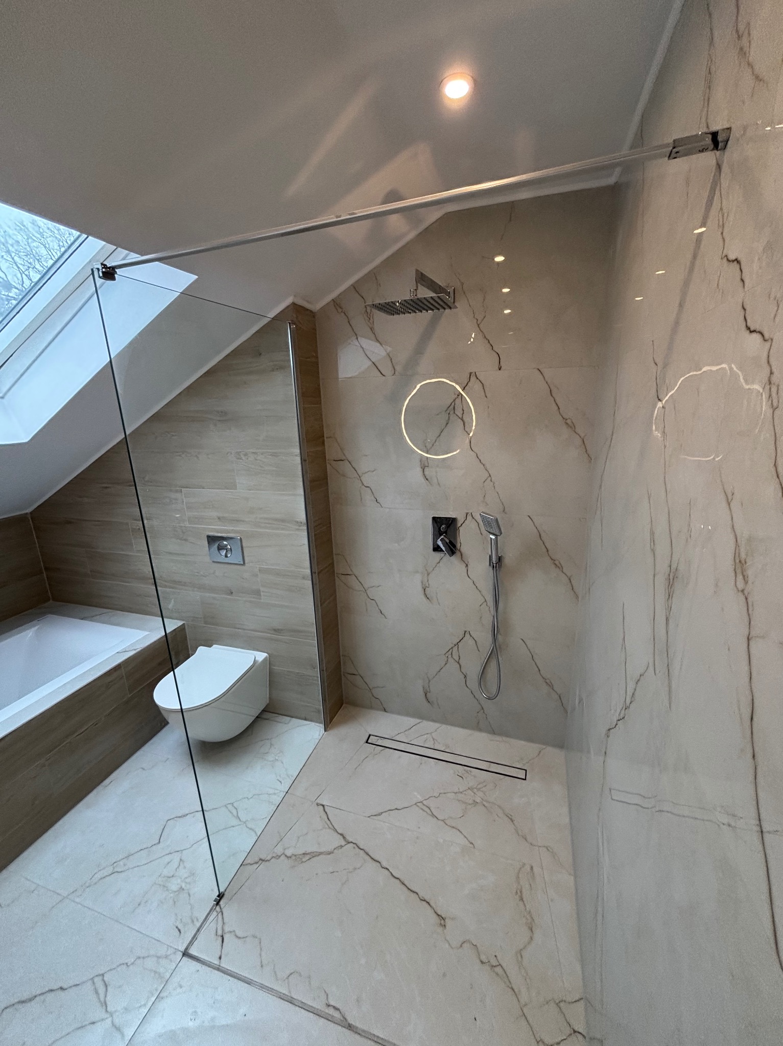 Wykończona łazienka na poddaszu z kabiną prysznicową typu walk-in, beżowe płytki imitujące marmur, toaleta podwieszana i okno dachowe. Nowoczesny design.