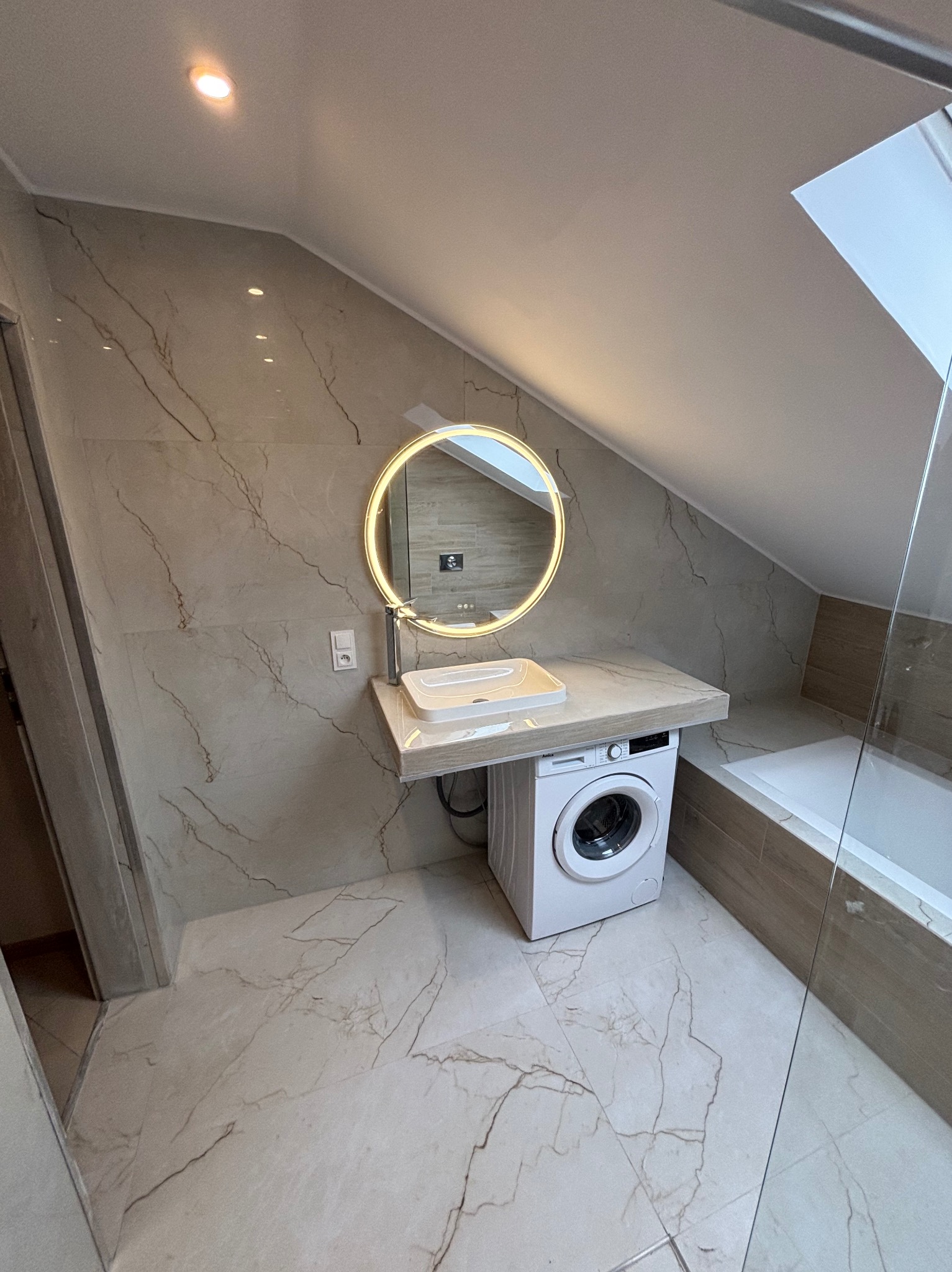 Jasna łazienka na poddaszu z marmurowymi płytkami, okrągłym lustrem z podświetleniem LED, umywalką nablatową i pralką pod blatem. Widoczna wanna za szklaną ścianką.