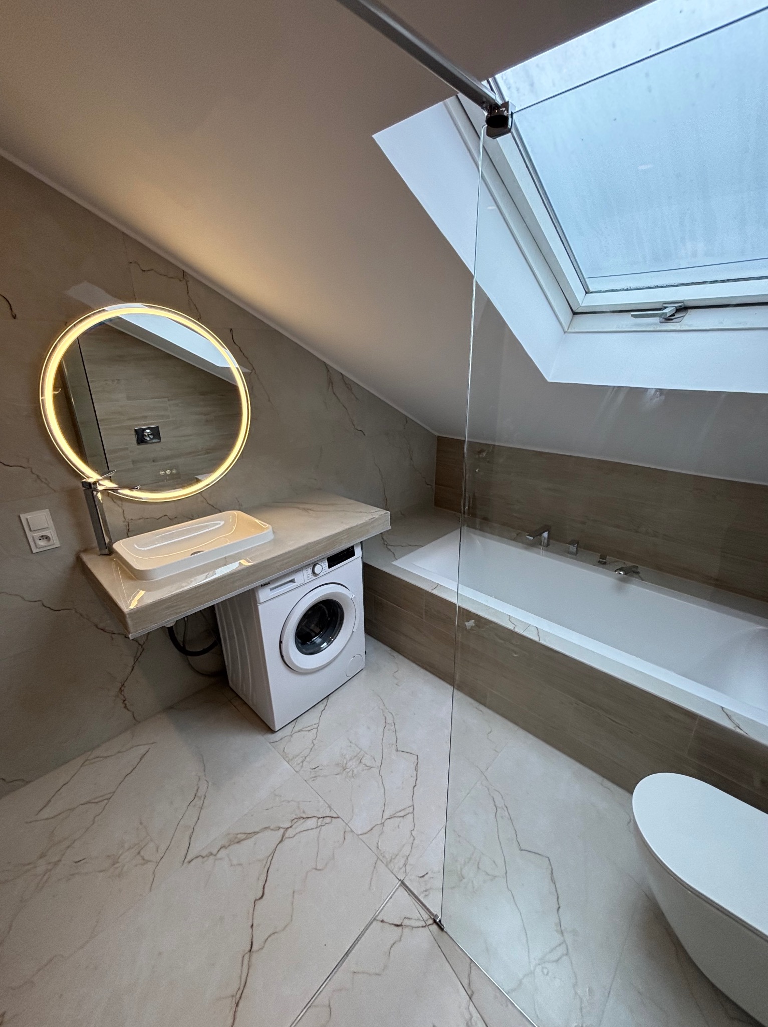 Jasna łazienka na poddaszu z oknem dachowym. Umywalka z podświetlanym lustrem nad pralką. Beżowe płytki imitujące marmur na podłodze i ścianach.