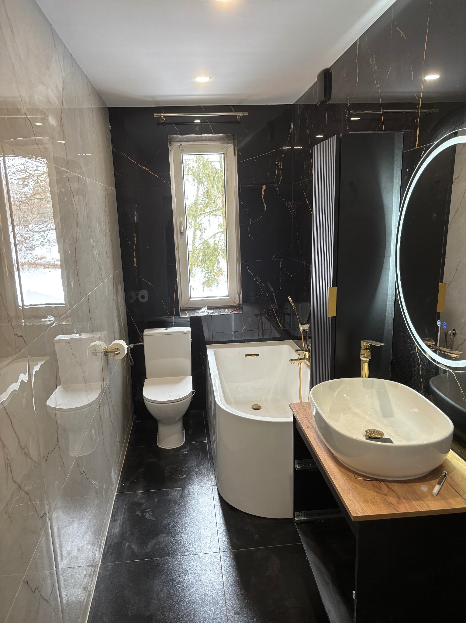 Elegancka łazienka po remoncie: czarne i marmurowe płytki, owalna umywalka na drewnianym blacie, wanna, toaleta i okrągłe lustro z podświetleniem LED.