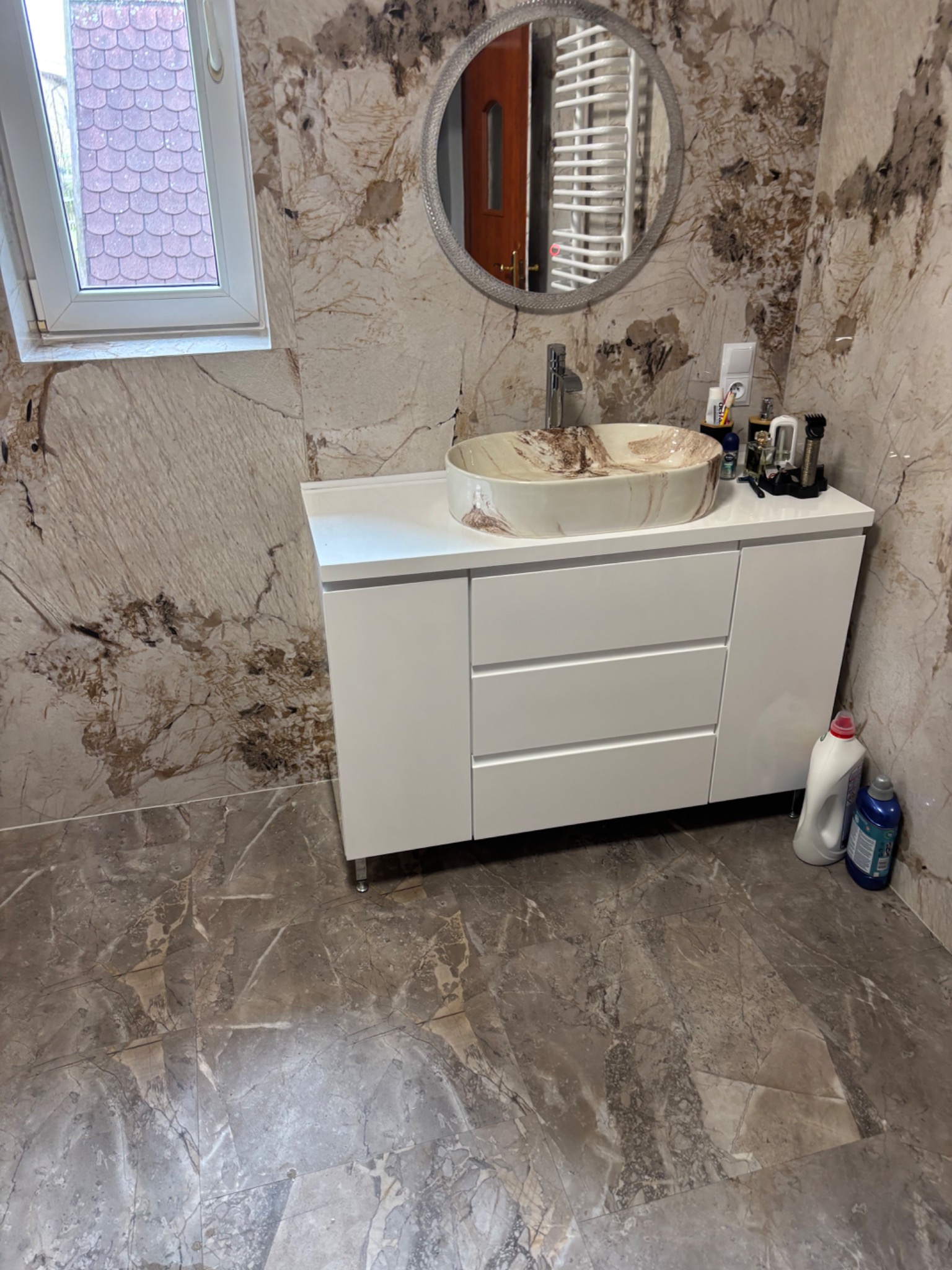 Wykończona łazienka z marmurowymi płytkami, okrągłym lustrem i białą szafką pod umywalką. Umywalka w odcieniach beżu i brązu. Widoczne akcesoria łazienkowe.