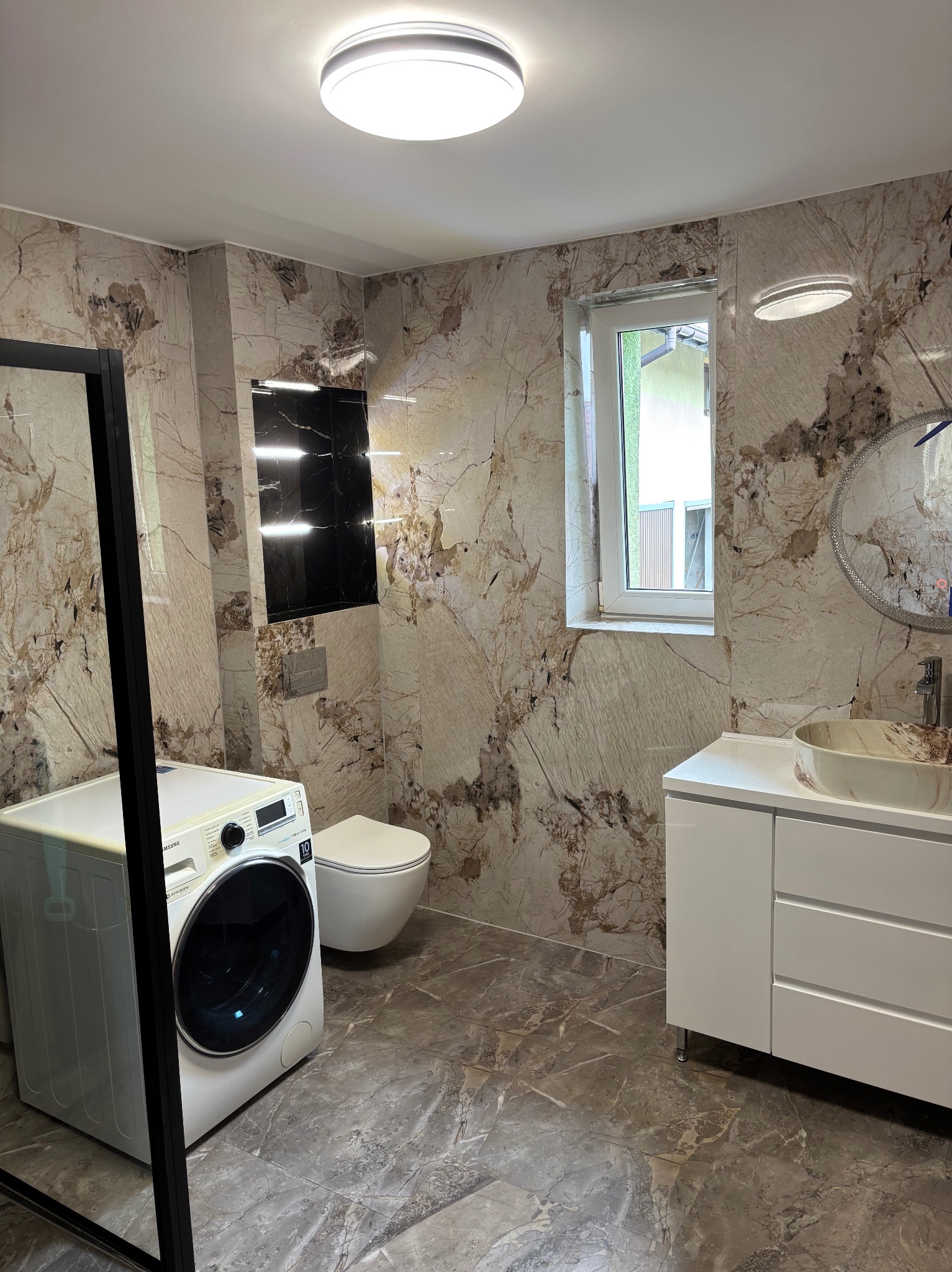 Elegancka łazienka z marmurowymi płytkami, umywalką nablatową i pralką. Czarna wnęka nad toaletą dodaje kontrastu. Okrągłe lustro i oświetlenie sufitowe.
