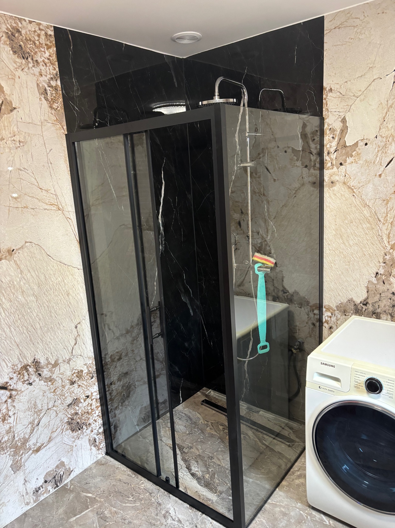 Nowoczesna kabina prysznicowa z czarną ramą i marmurowymi płytkami w łazience. Widoczna pralka i akcesoria prysznicowe.