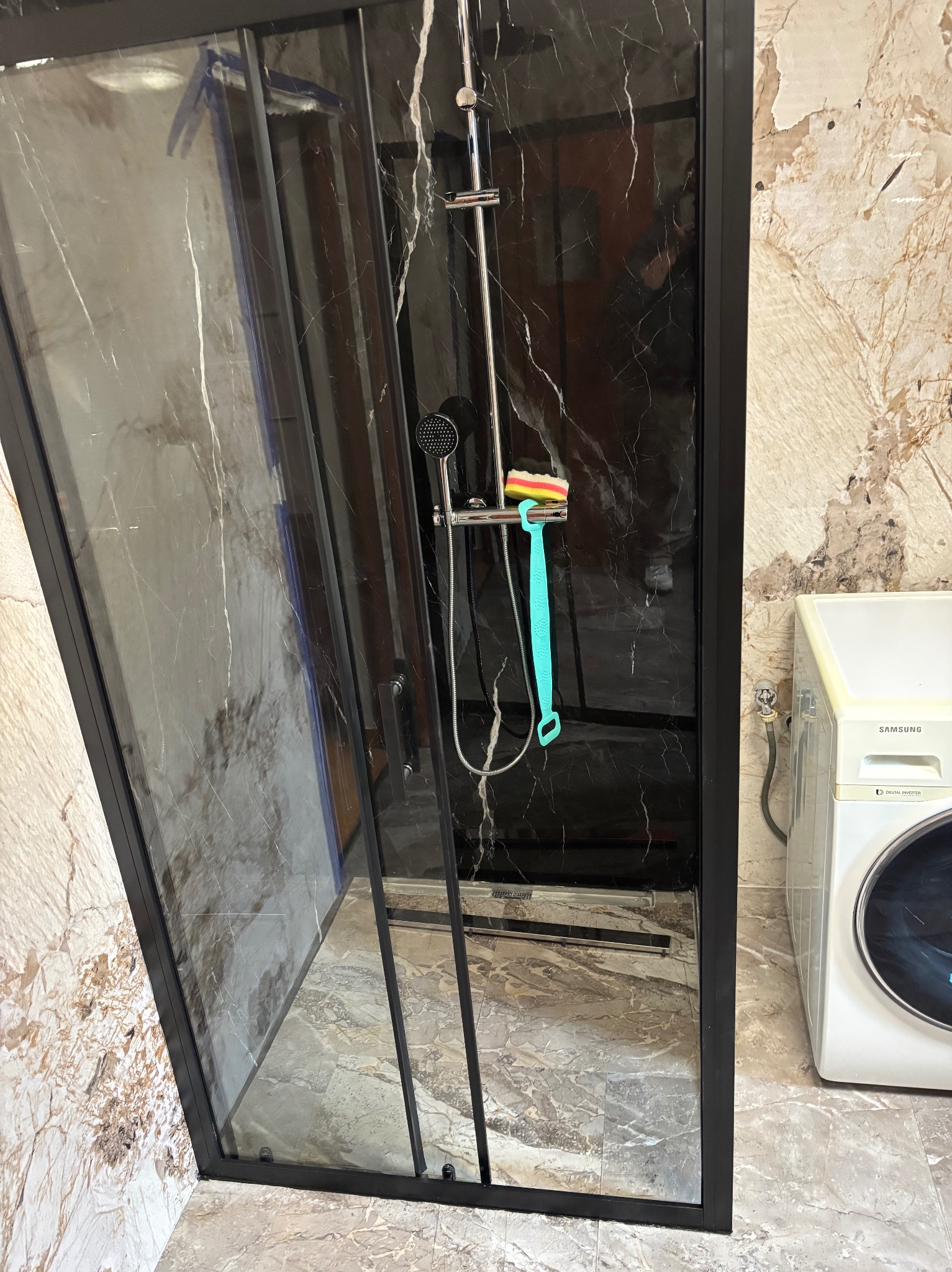 Nowoczesna kabina prysznicowa z czarnymi ramami i marmurowym wykończeniem, widoczna obok pralka. Minimalistyczny design, eleganckie detale i jasne, odbijające światło powierzchnie.