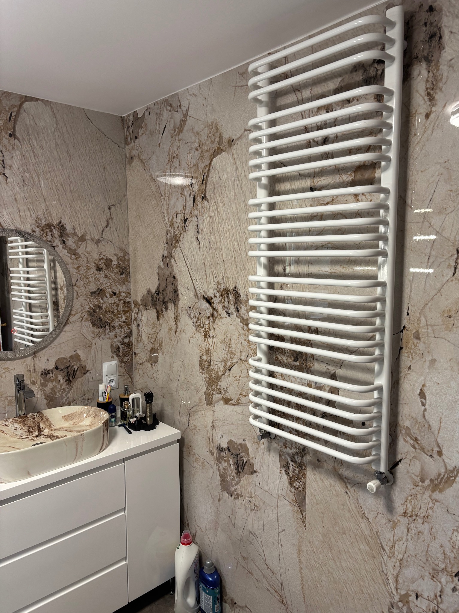 Wykończona łazienka z marmurowymi płytkami, białą szafką pod umywalką i grzejnikiem drabinkowym. Umywalka w odcieniach beżu i brązu. Nowoczesny design.