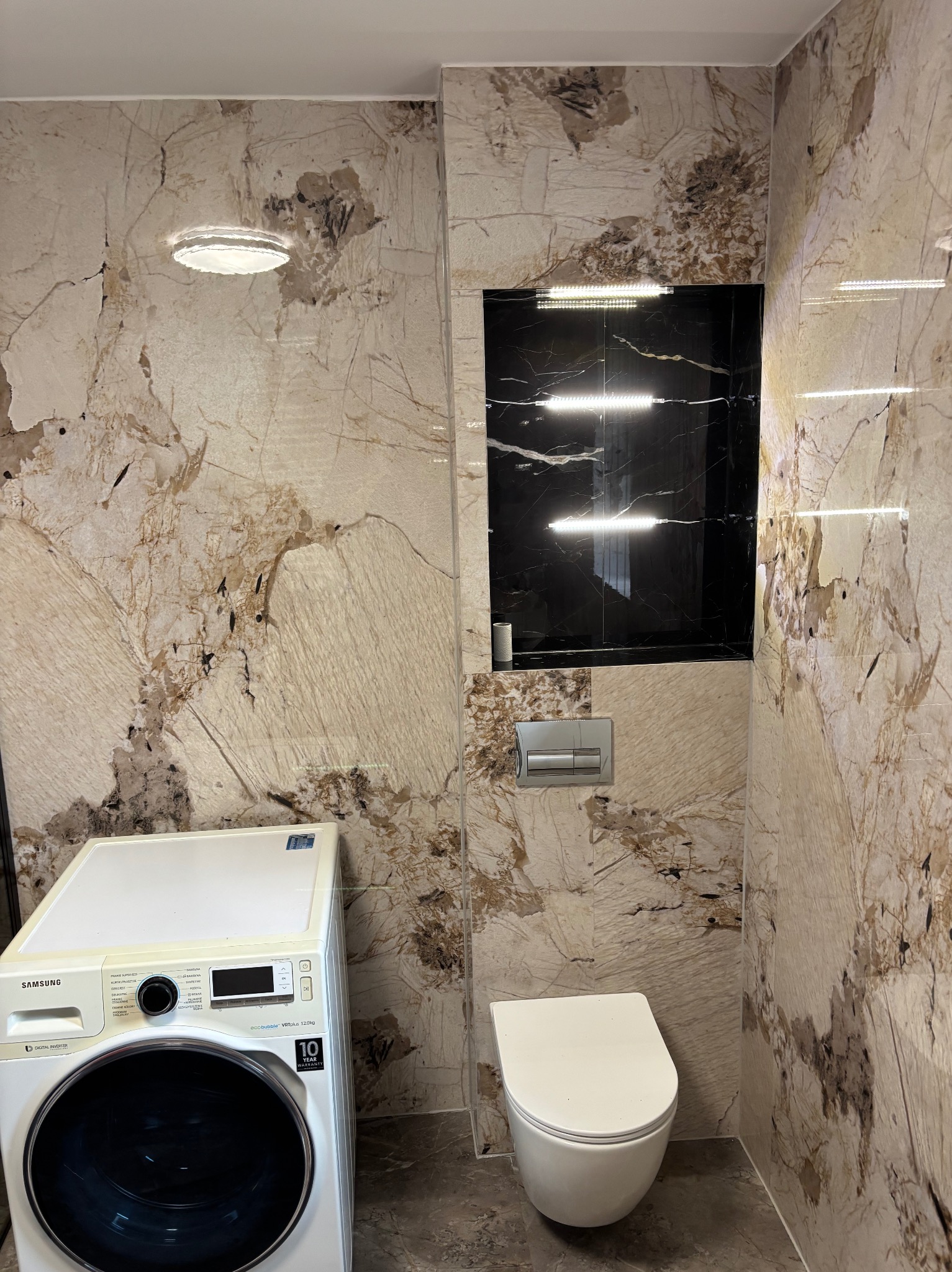 Wykończona łazienka z płytkami imitującymi marmur, podświetlaną wnęką nad toaletą i pralką. Nowoczesny design z akcentami beżu i czerni.