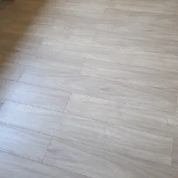 Jasne, drewnopodobne panele ułożone w jodełkę na podłodze, widoczne przy listwie przypodłogowej z gniazdkiem elektrycznym w rogu pomieszczenia.