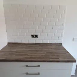 Ściana nad blatem kuchennym wyłożona białymi, wypukłymi płytkami typu metro, z widocznymi gniazdkami elektrycznymi, blat drewnopodobny, szafki dolne z trzema szufladami i metalowymi uchwytami.