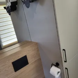 Nowoczesna łazienka z drewnopodobnymi płytkami, zabudową meblową w kolorze jasnego beżu i oryginalnymi uchwytami na papier toaletowy w kształcie owiec.