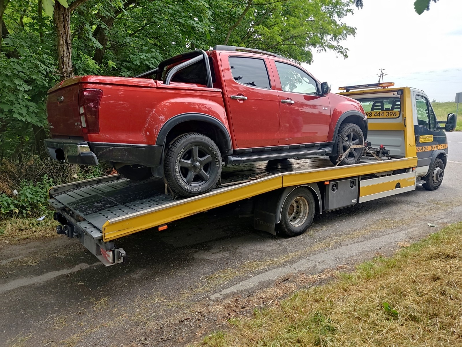 Czerwony pickup Isuzu D-Max zabezpieczony pasami na żółtej platformie lawety pomocy drogowej w trakcie transportu, widok z boku na tle zieleni.