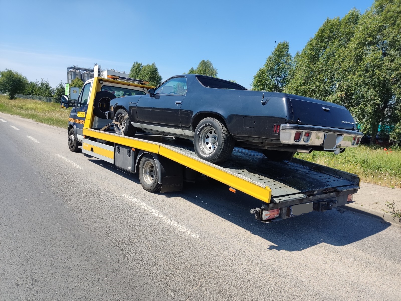 Czarny klasyczny samochód typu El Camino zabezpieczony pasami na żółtej platformie lawety, widok z tyłu i boku, na tle drogi i zieleni.