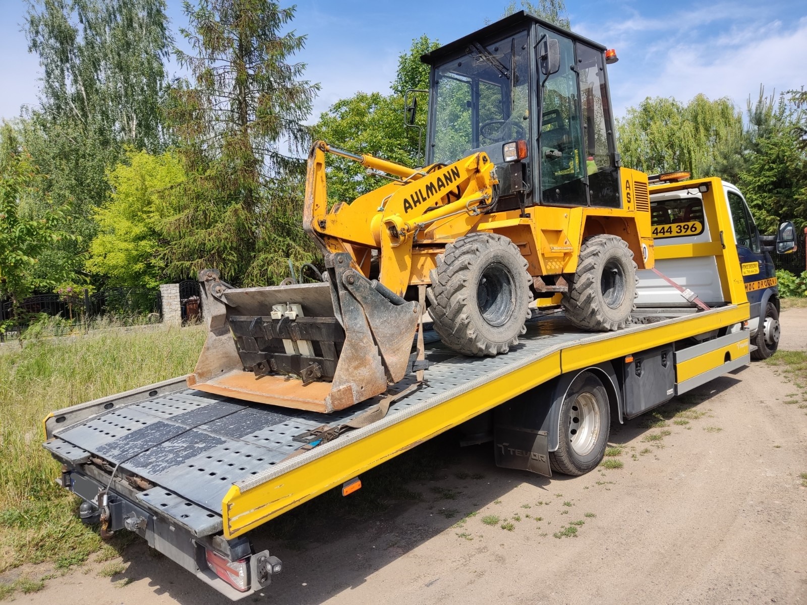 Żółta ładowarka Ahlmann, zabezpieczona pasami, na platformie lawety w słoneczny dzień.