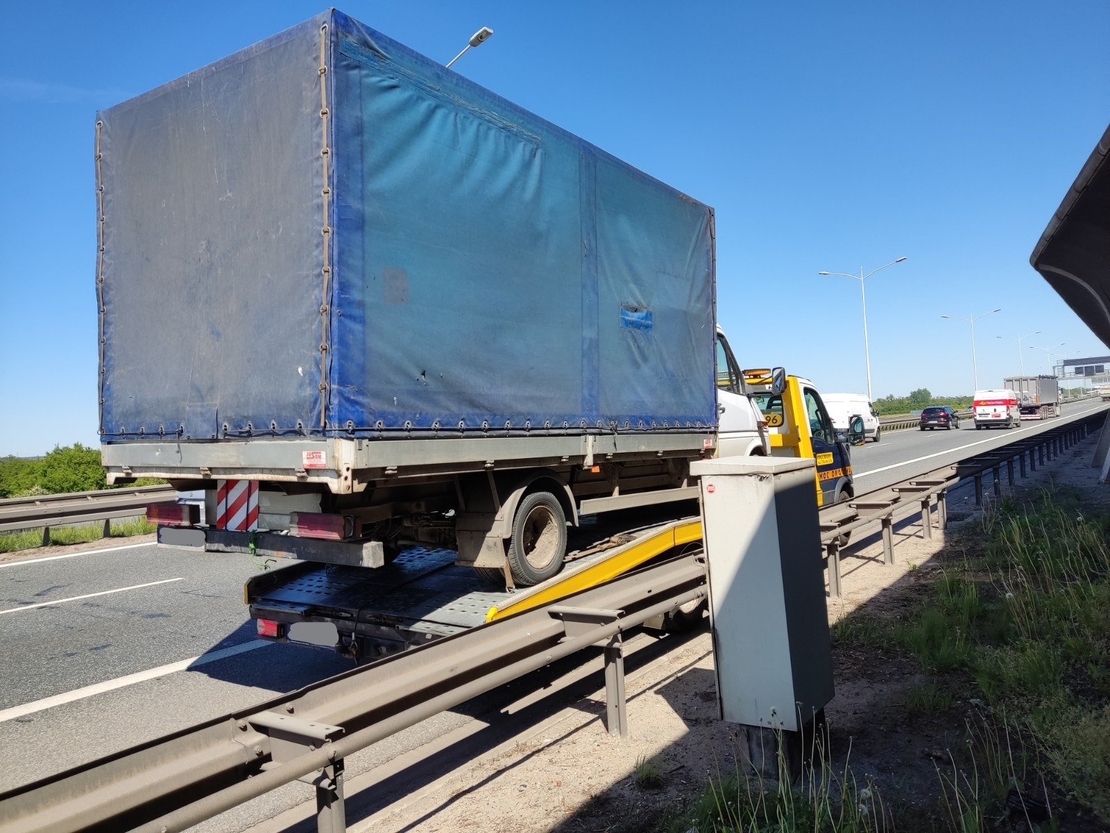 Ciężarowy samochód z plandeką, z uszkodzonym kołem, transportowany na lawecie na autostradzie w słoneczny dzień.