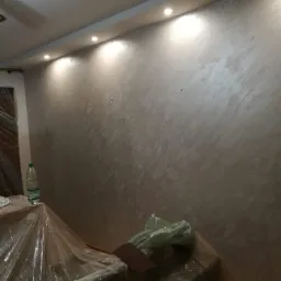 Ściana w pokoju po nałożeniu dekoracyjnej, beżowo-srebrnej gładzi, oświetlona punktowymi lampami sufitowymi. Meble zabezpieczone folią malarską.