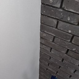 Beffsan - Fragment ściany z białym tynkiem obok fragmentu ściany wykończonej ciemnoszarą cegłą licową, widoczny fragment rusztowania.