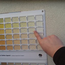 Beffsan - Wzornik tynków Caparol z paletą odcieni beżu i brązu, palec wskazuje na odcień 'Mais 18' na tle ściany o podobnej fakturze.