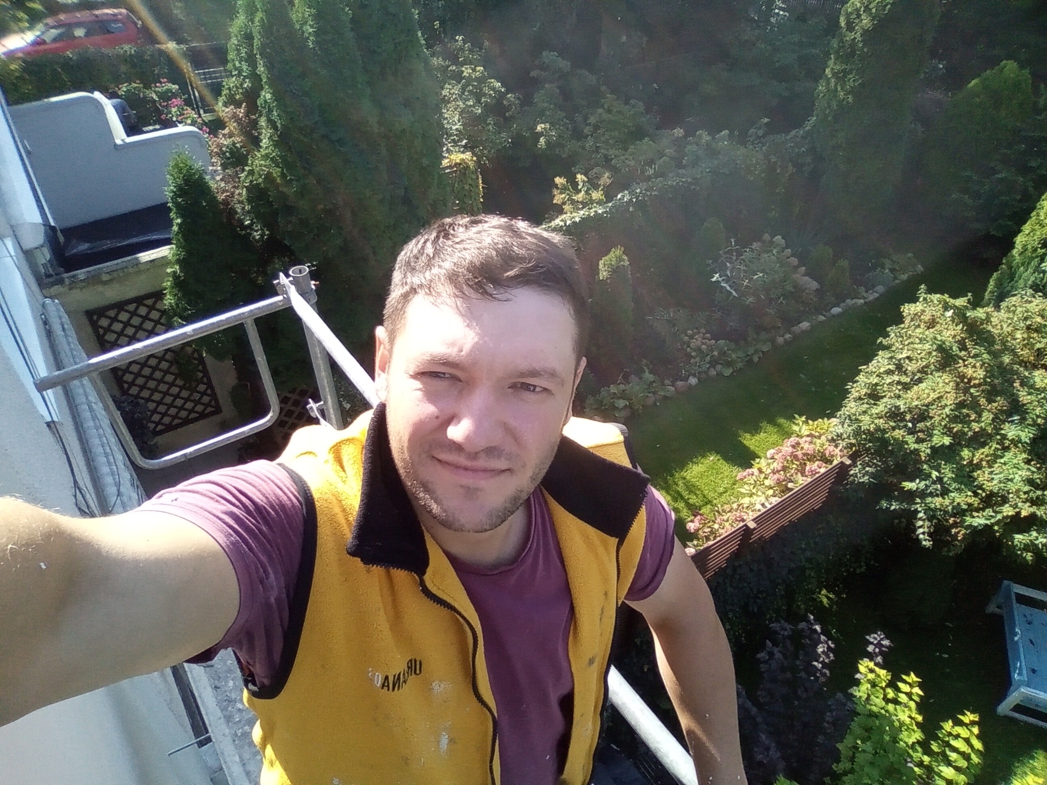 Mężczyzna w żółtej kamizelce roboczej robiący selfie na tle zielonego ogrodu z balkonu podczas prac na wysokości.