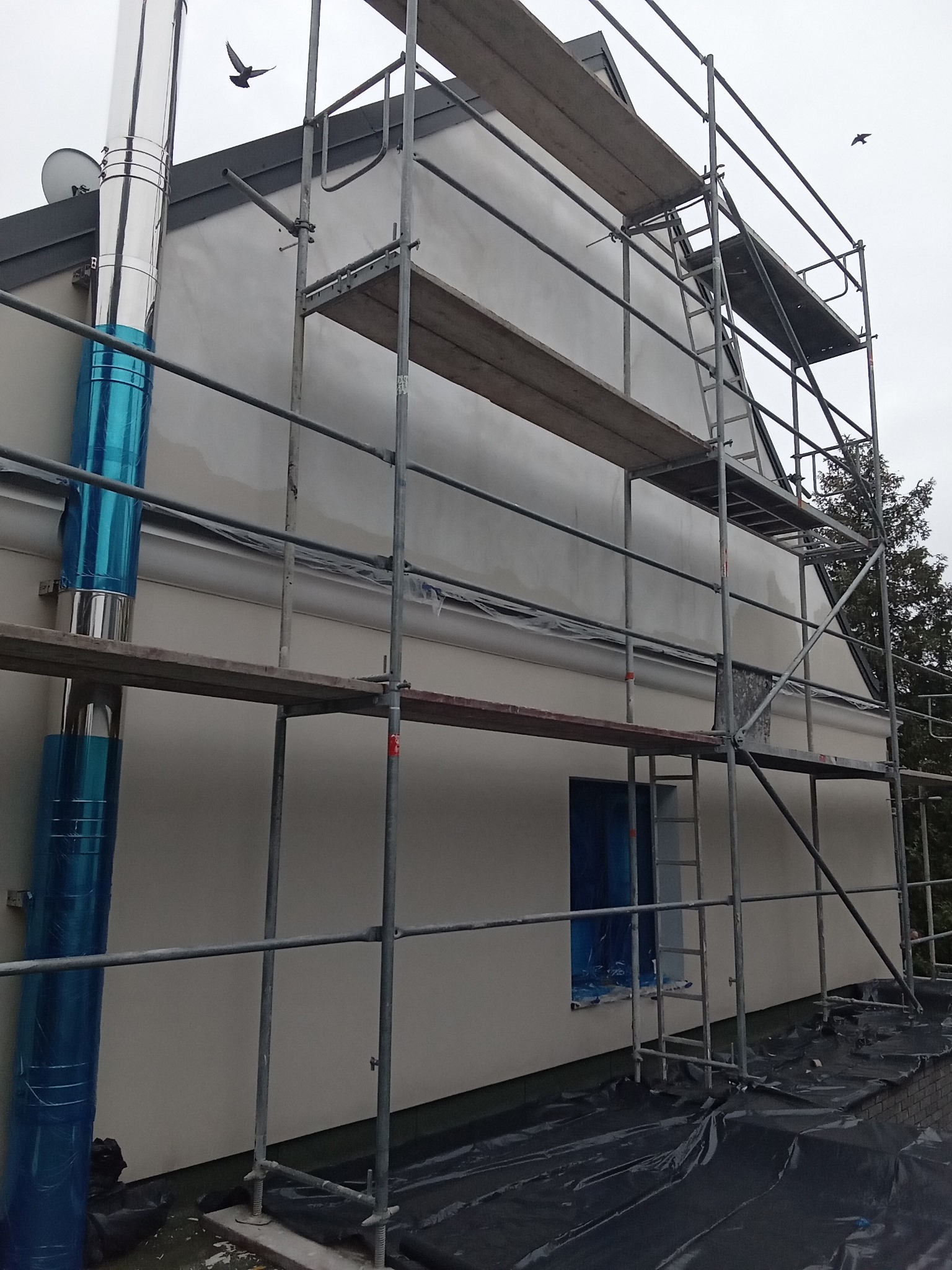 Dom jednorodzinny w trakcie prac elewacyjnych, widoczne rusztowania, zabezpieczone okno folią, świeżo nałożona warstwa szpachli, komin w niebieskiej folii ochronnej, ptak w locie.