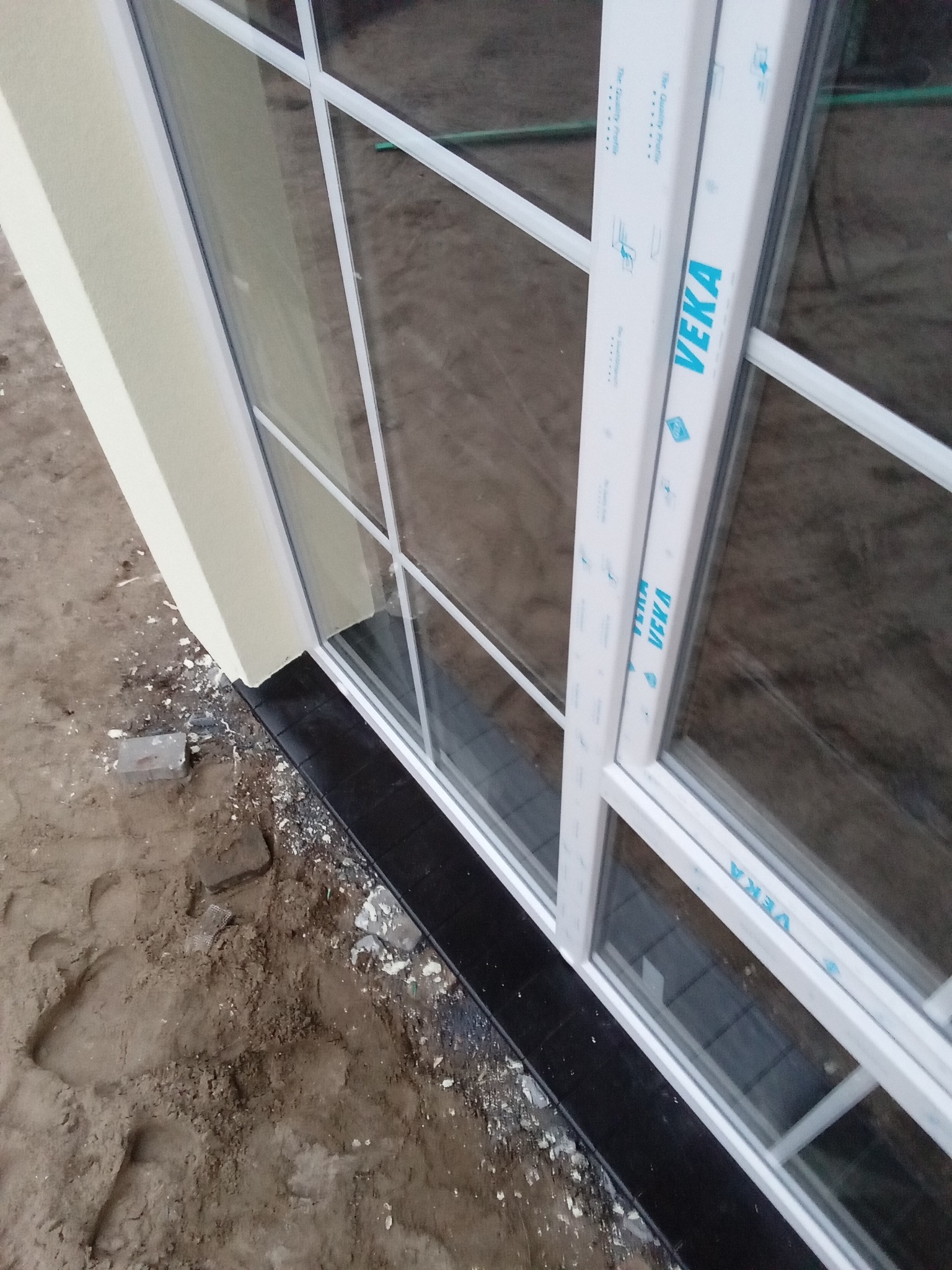 Fragment ściany budynku w Warszawie z ociepleniem i zamontowanym oknem PCV marki Veka, widoczne ślady prac budowlanych na ziemi.