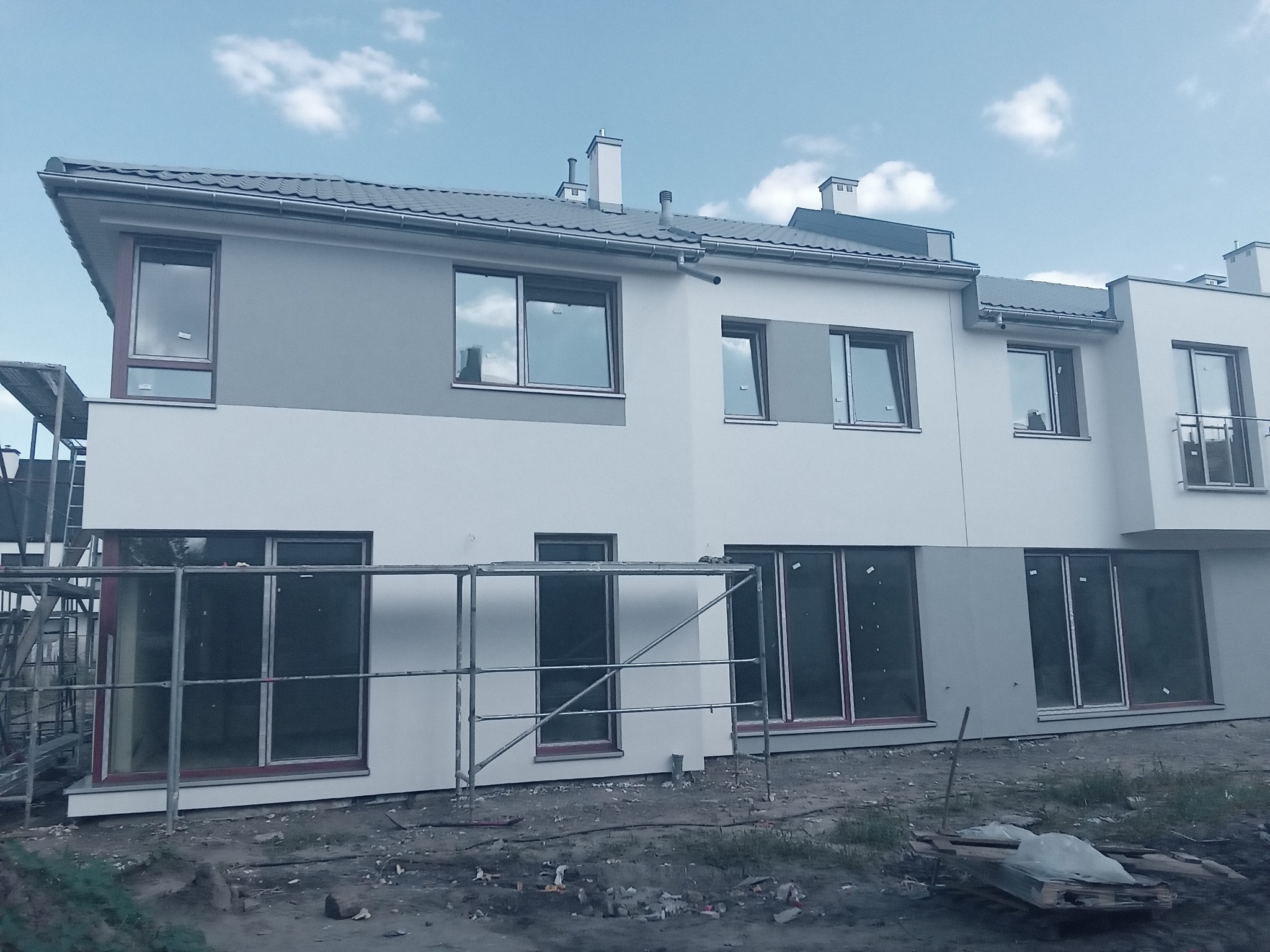 Nowo wybudowany dom szeregowy z szarą i białą elewacją, częściowo zakryty rusztowaniami, z widocznymi oknami i elementami wykończeniowymi dachu.
