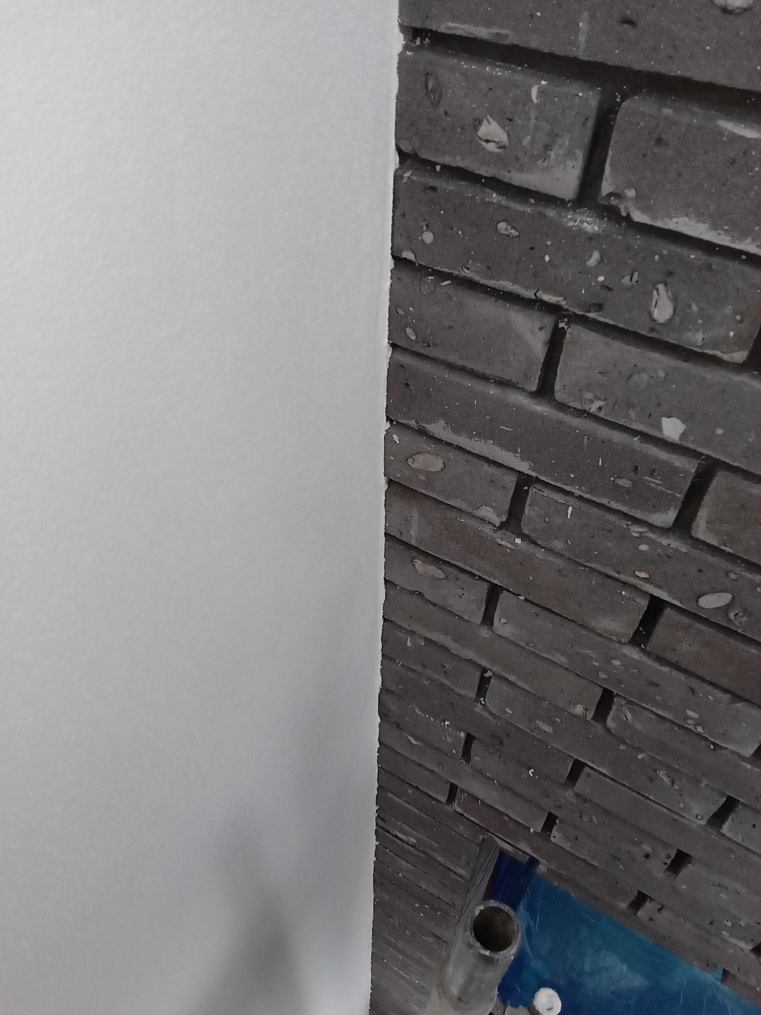 Fragment ściany z białym tynkiem obok fragmentu ściany wykończonej ciemnoszarą cegłą licową, widoczny fragment rusztowania.