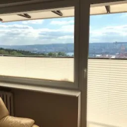 Białe, plisowane osłony okienne zamontowane w oknach z widokiem na panoramę miasta z elementami portowymi i zielenią w tle.