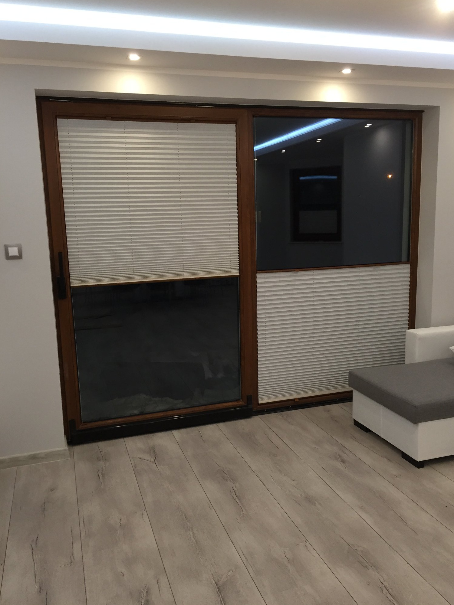 Zamknięte, jasne plisy w brązowych ramach okiennych na dwuskrzydłowych drzwiach balkonowych w Kościerzynie, widoczne fragmenty białej sofy i podłogi imitującej drewno.