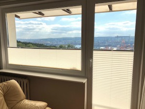 Białe, plisowane osłony okienne zamontowane w oknach z widokiem na panoramę miasta z elementami portowymi i zielenią w tle.